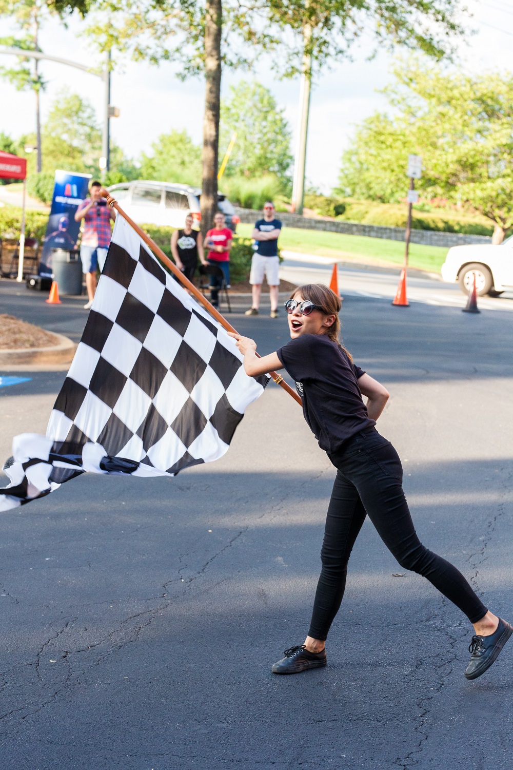 Girl holding a checkered flag in a parking lot with people and trees in background.