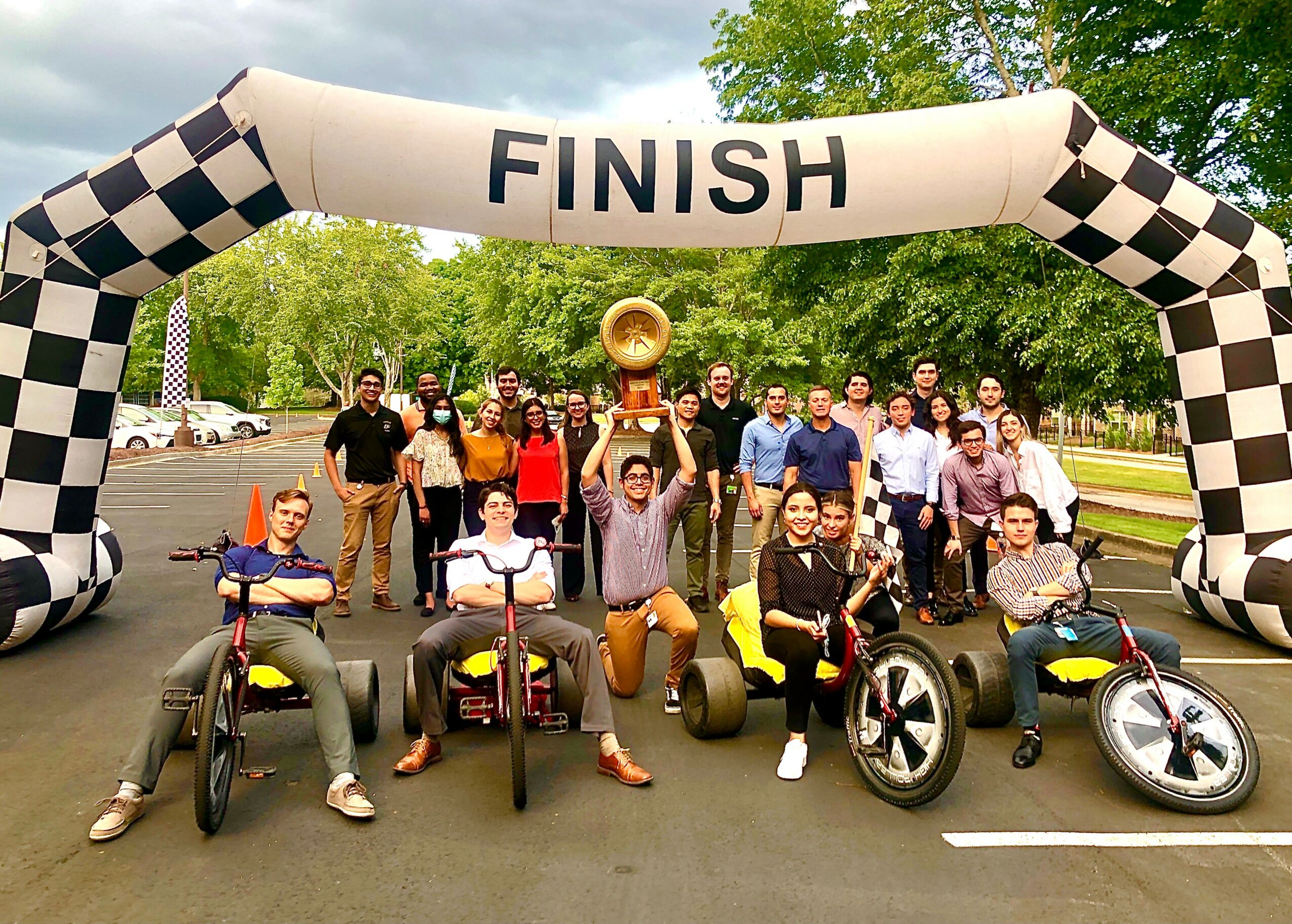 Group of people celebrating at a race finish line with go-karts and a person holding a trophy, outdoors with trees.