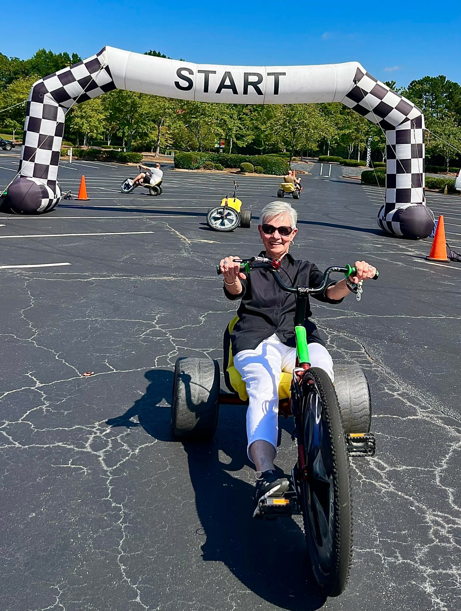 Woman riding a tricycle in a parking lot under a start banner, with trees and orange cones in the background.