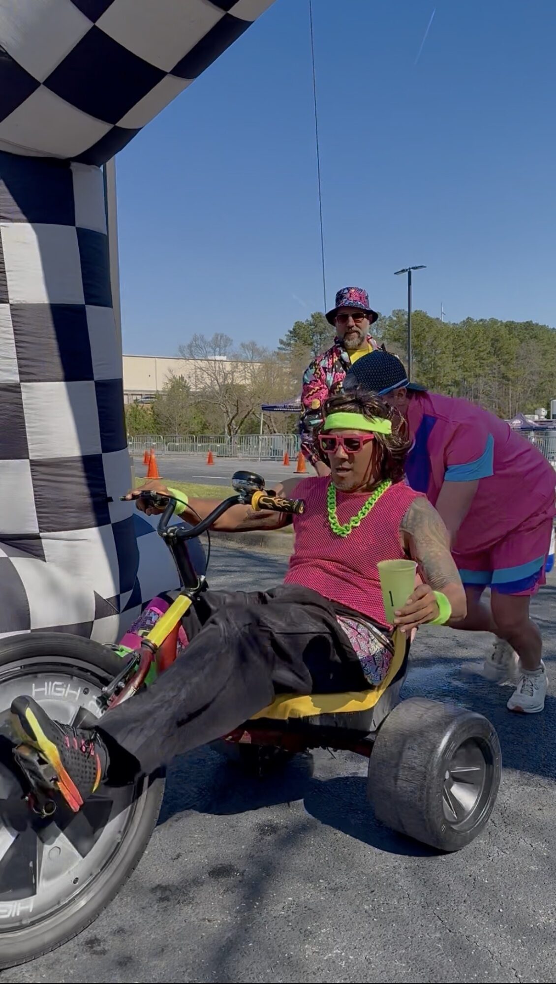 Person in pink shirt and sunglasses sitting on a tricycle, with two other people nearby, outdoors on a sunny day.