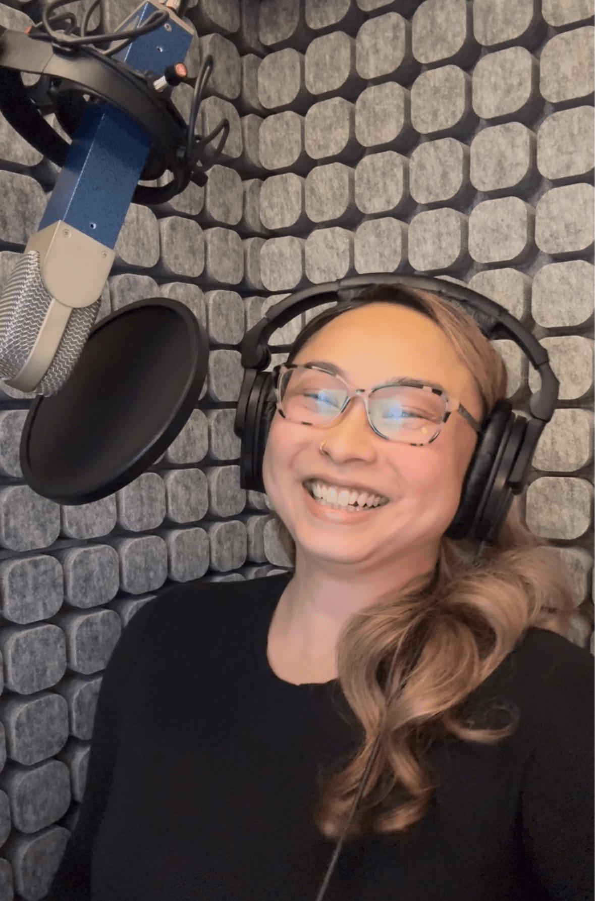 Woman with glasses smiling in a recording studio with a microphone and soundproof wall background.