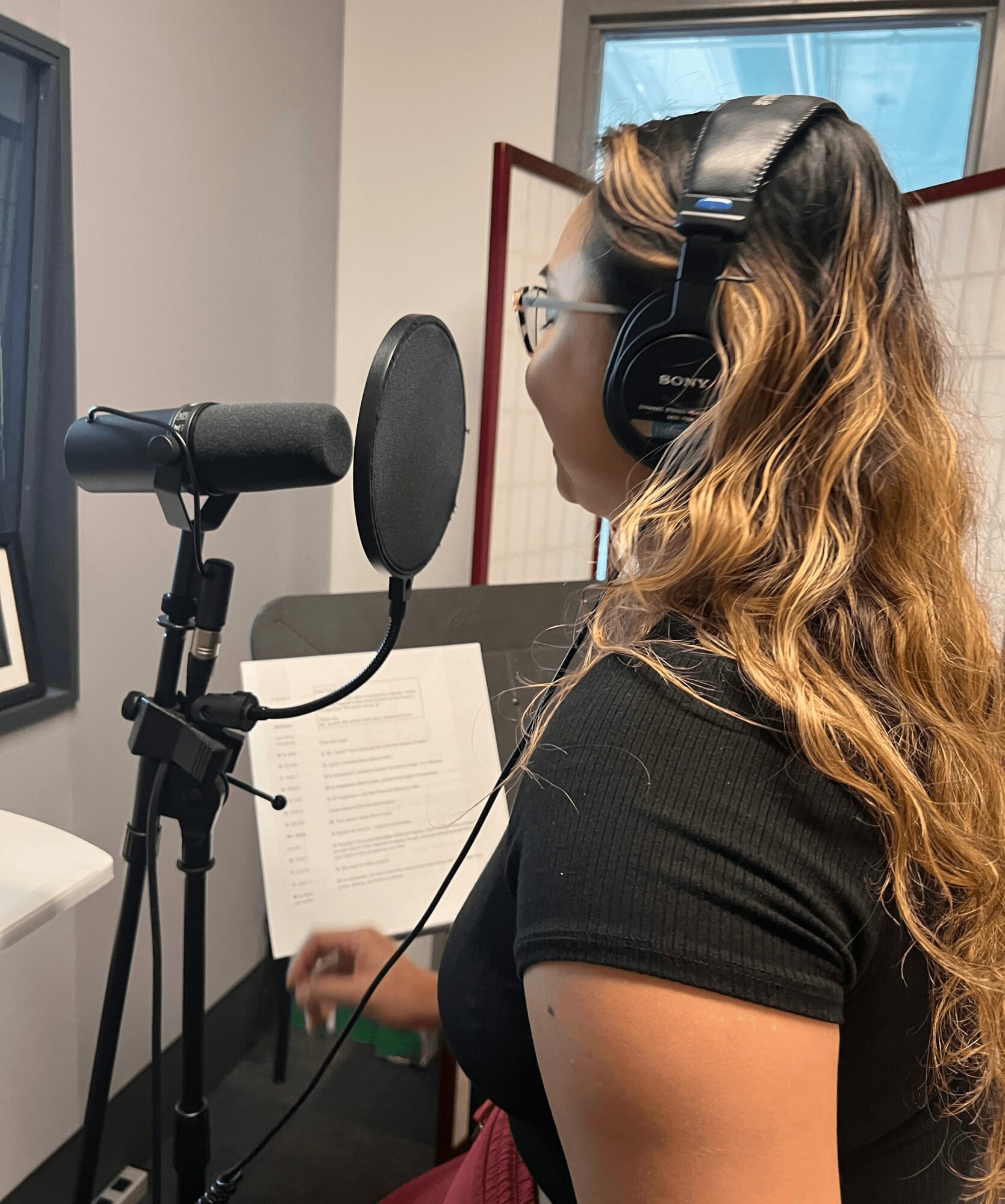Woman with headphones singing into a microphone in a recording studio, reading from a music stand.