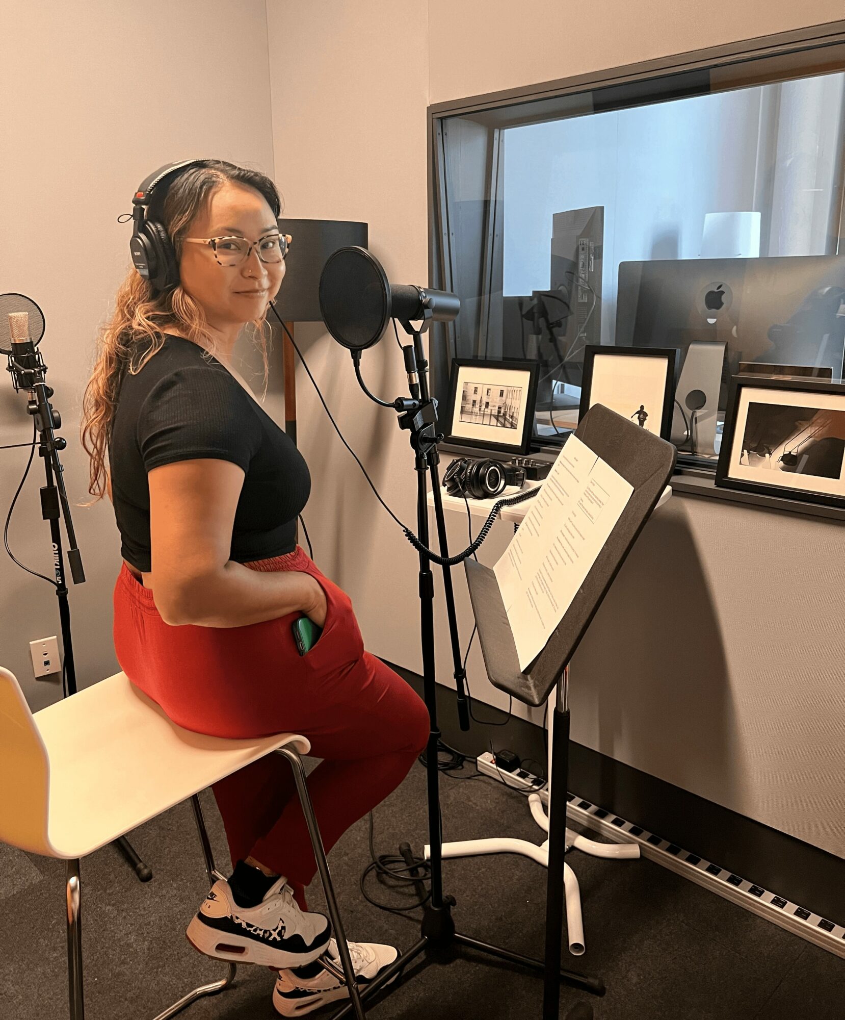 Woman with headphones sitting in recording studio, microphone, music stand, and framed pictures on window sill.