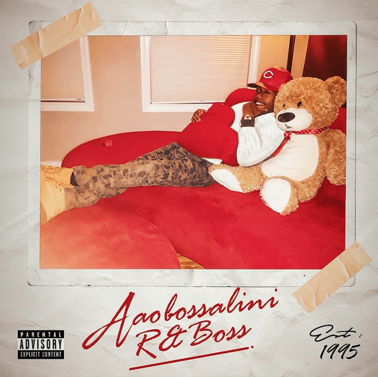 Person lying on a red couch with a large teddy bear, wearing a red cap and patterned pants, in a room with window blinds.