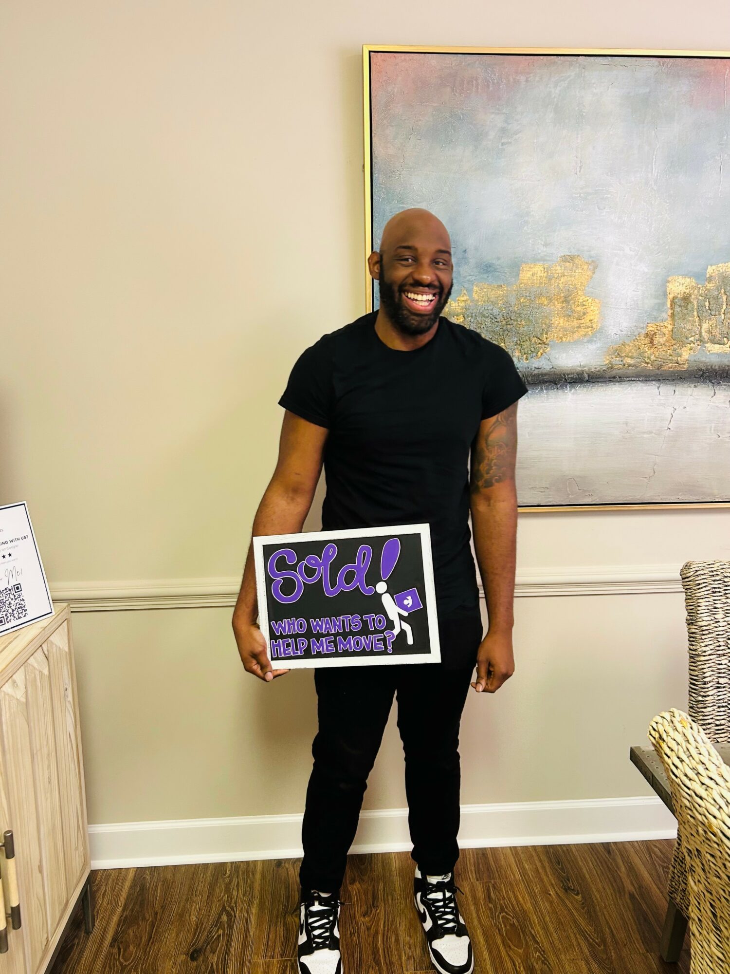 Man with a beard smiling, holding a framed sign, standing in a room with a large painting on the wall.