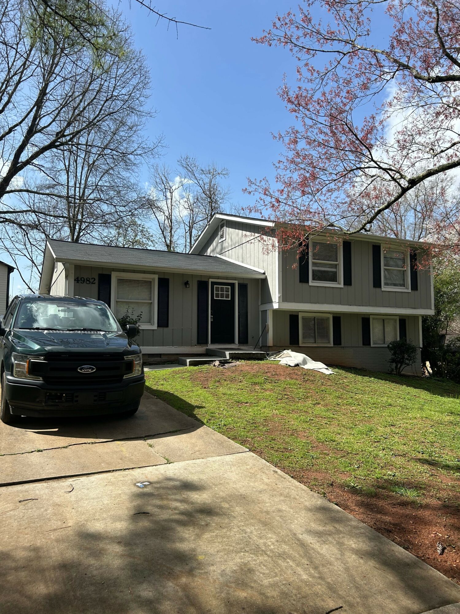 Two-story house with a porch, parked car in driveway, trees, and a sidewalk, viewed from the side.