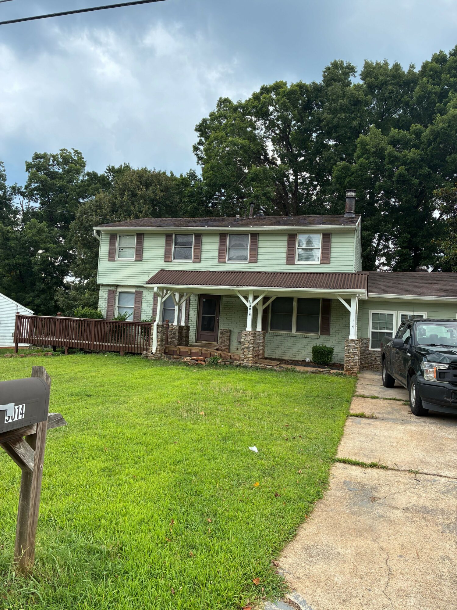 Two-story house with a porch, surrounded by trees and a green lawn, with a driveway and parked vehicle nearby.