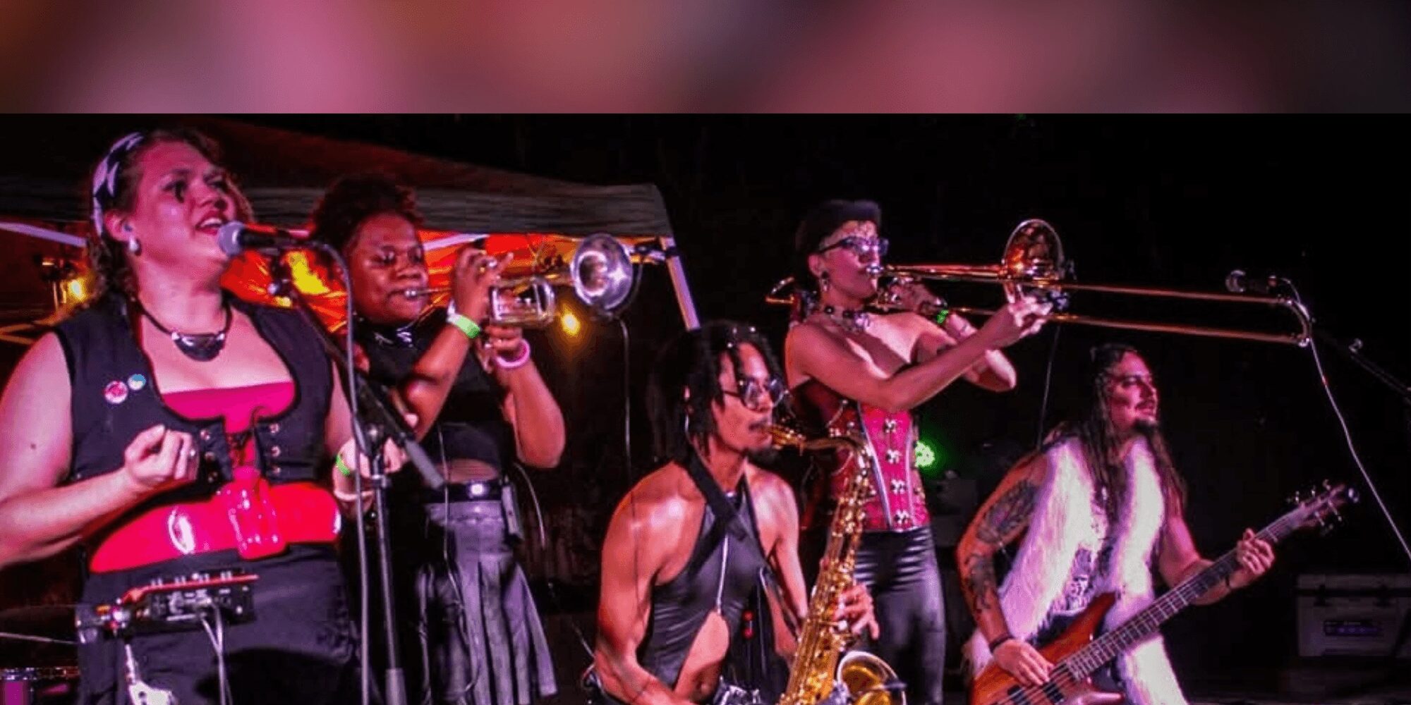 Group of five women performing on stage with musical instruments, singing and playing trumpet and guitar, in a dark setting.
