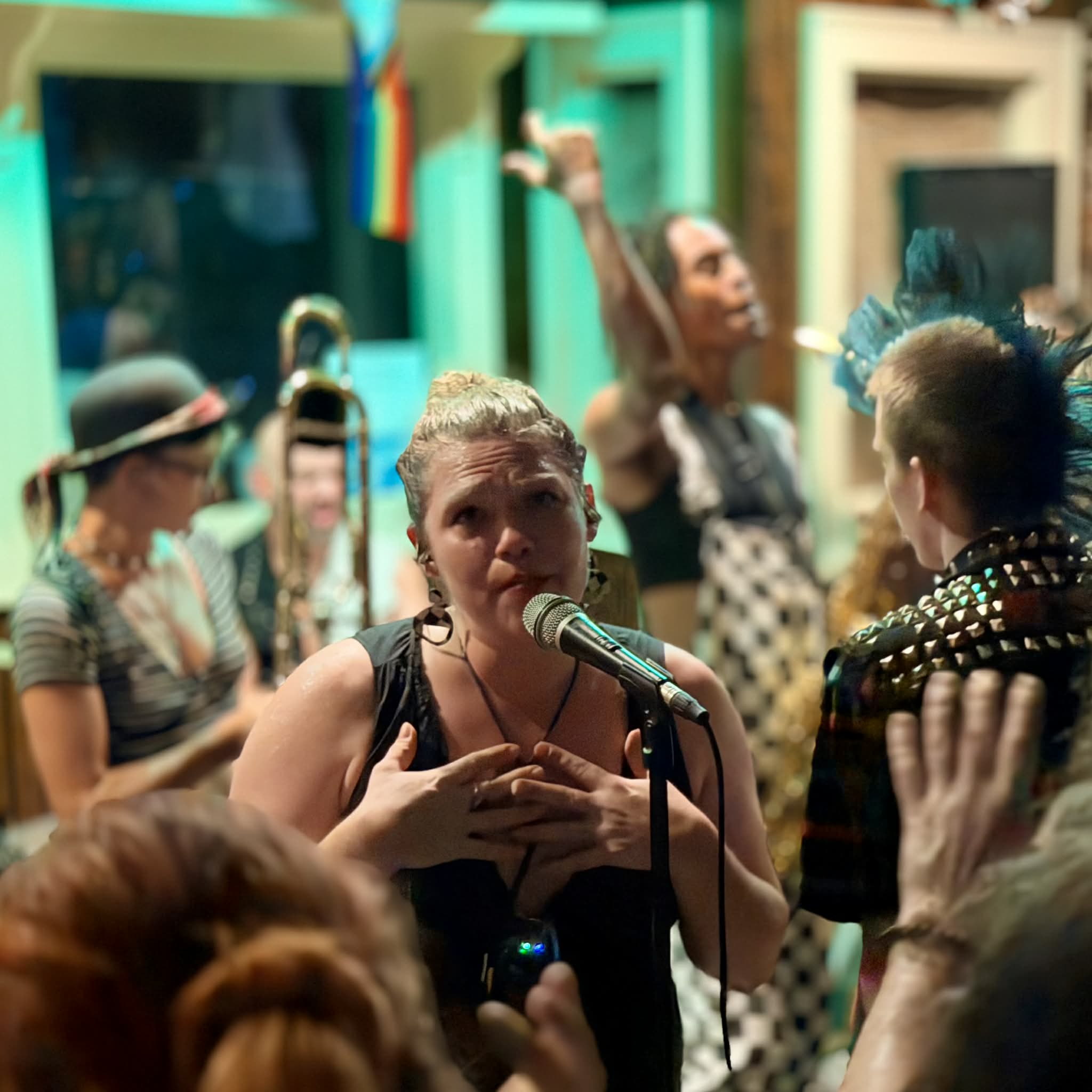 Woman speaking into microphone with hands on chest, surrounded by people in colorful clothing and accessories, lively setting.