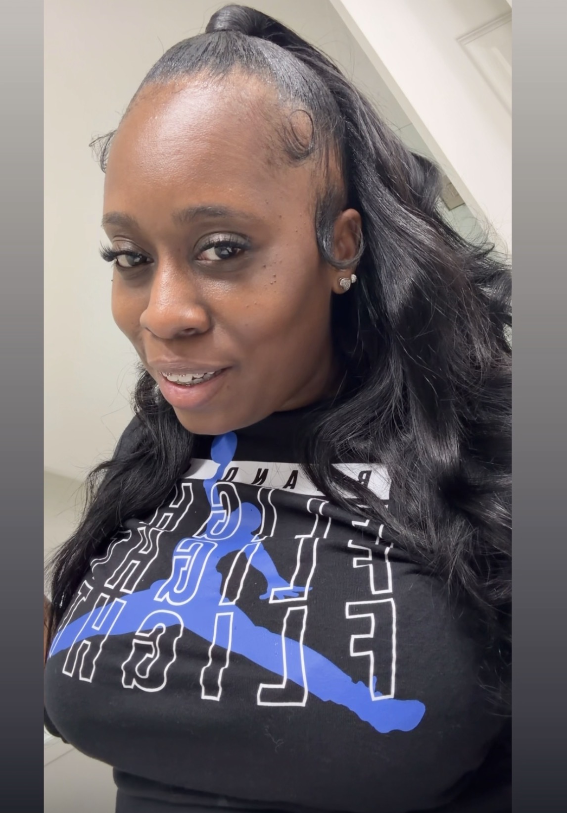 Woman with long, wavy hair in a high ponytail, smiling, wearing a black shirt with blue and white design, indoors.