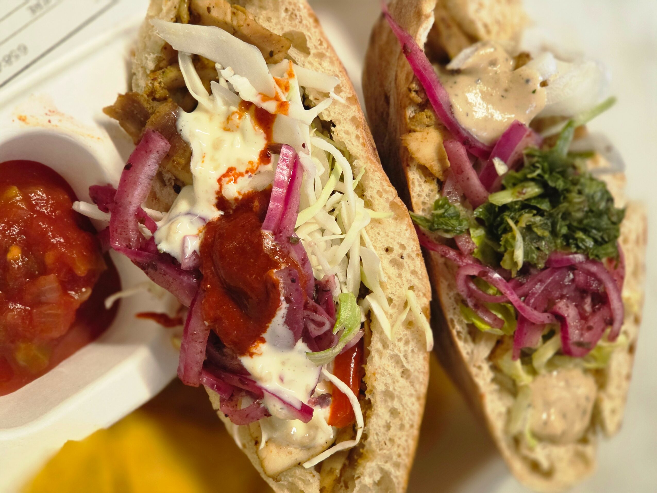 Two sandwiches with bread, shredded cheese, vegetables, and sauces, served with a side of salsa or chutney.