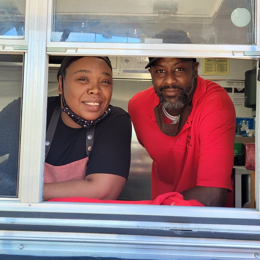 Two people leaning out of a food truck window, smiling at the camera, with a kitchen in the background.