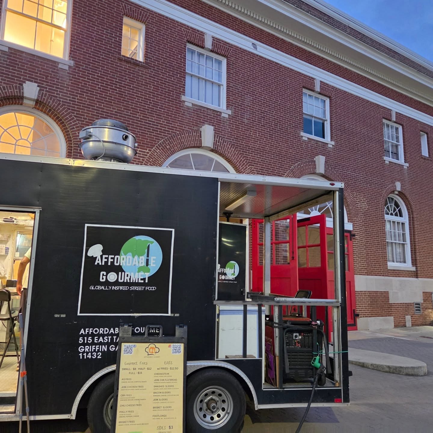 Food truck parked on street in front of brick building with arched windows, serving area with red equipment, menu displayed outside.
