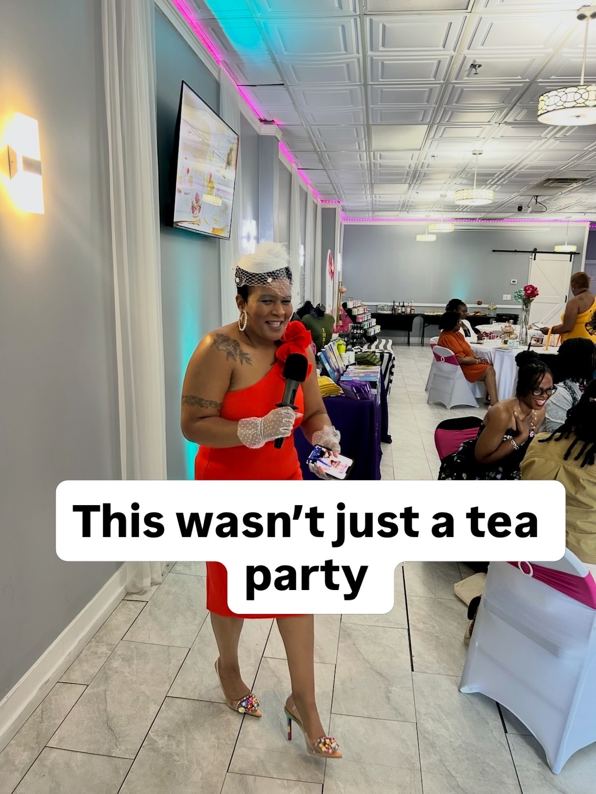 Woman in red dress holding a microphone at a tea party event, with guests seated at tables in the background.
