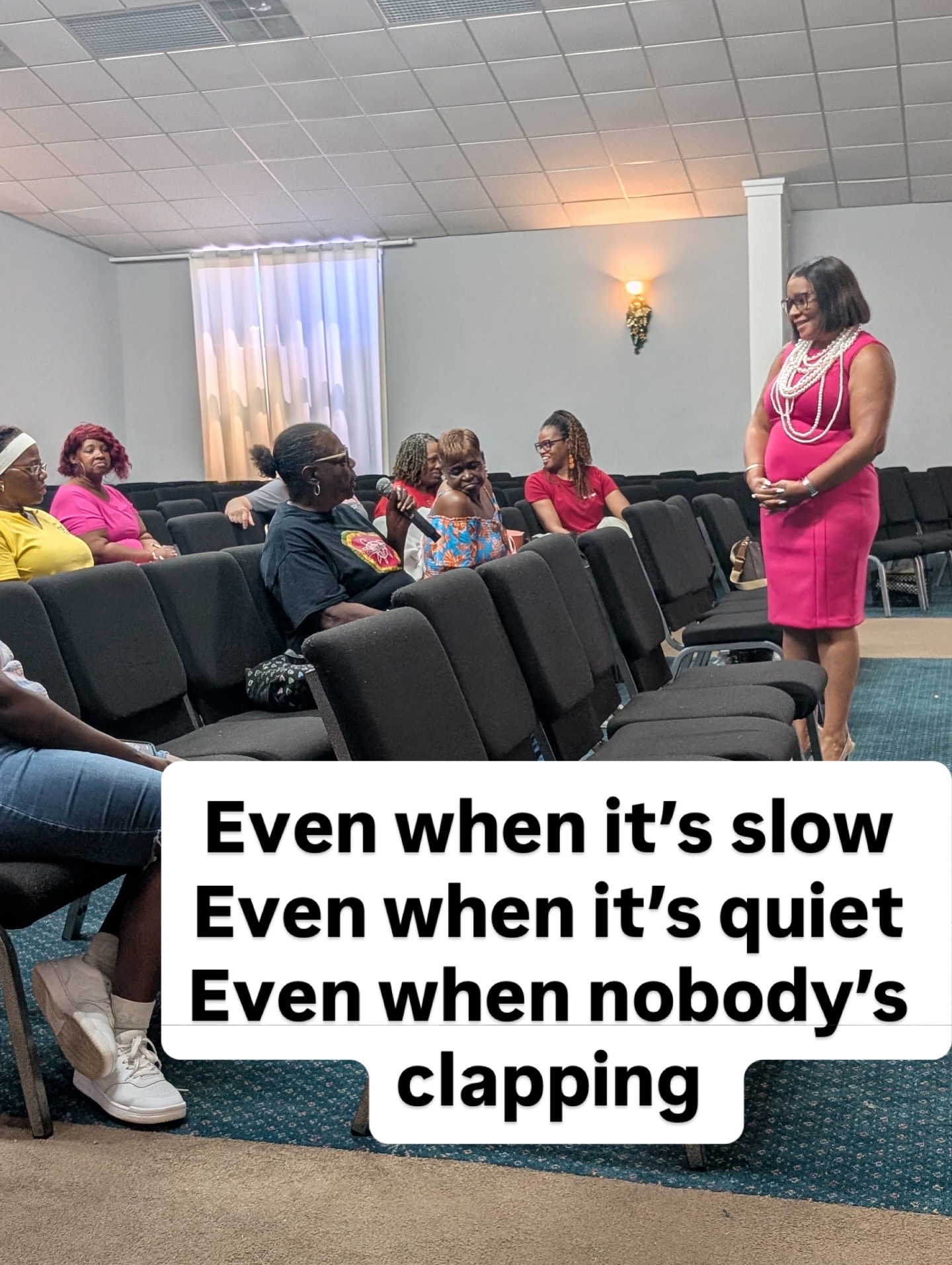 A woman in a pink dress stands in front of seated people in a room with a light-colored wall and window, speaking to the group.