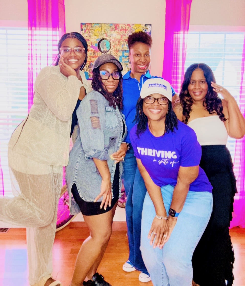 Five women smiling indoors, colorful curtains and artwork in the background, one wearing a blue shirt with 'THRIVING' text.