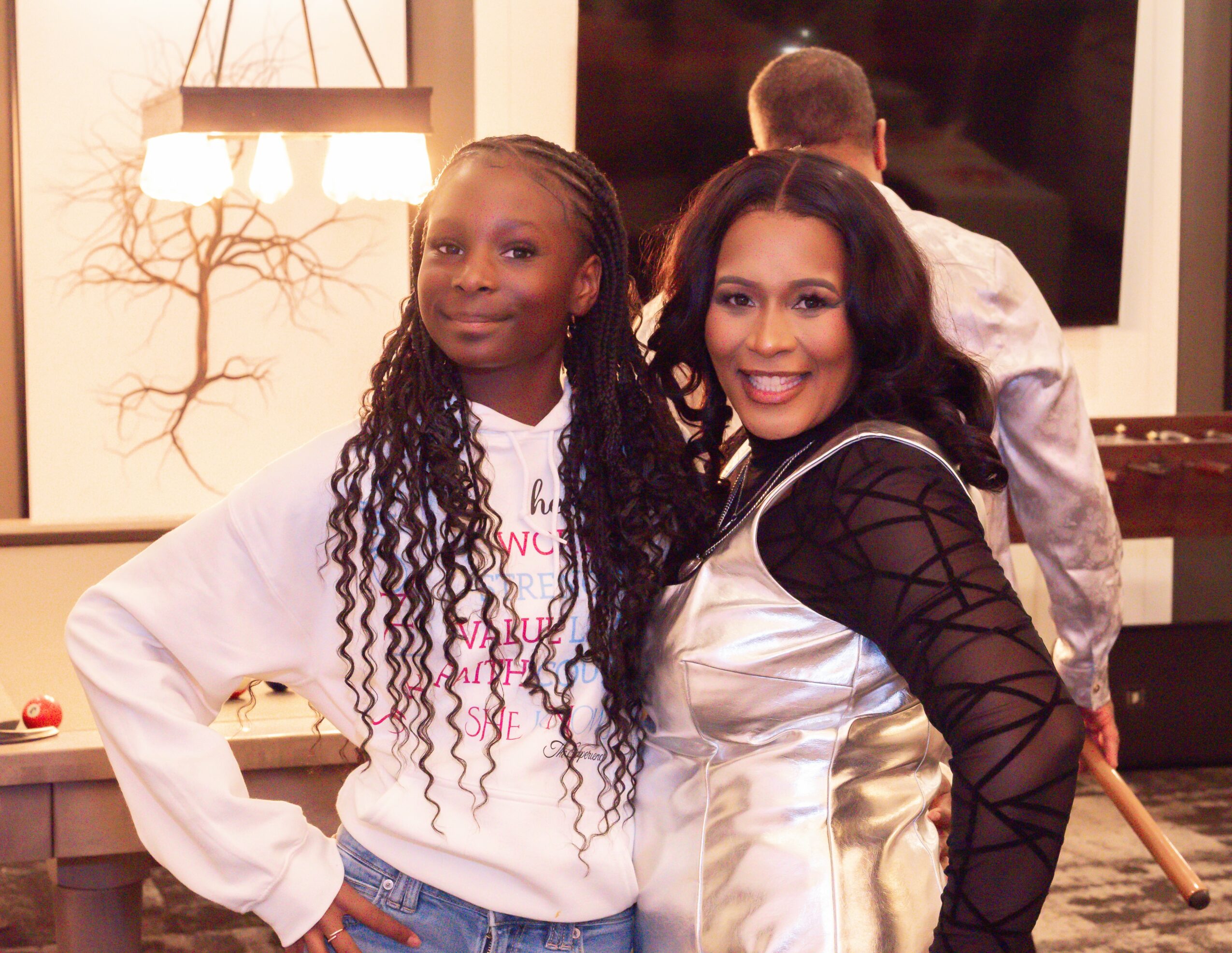 Two women smiling indoors, one with long curly hair and the other with shoulder-length dark hair, in front of a television.