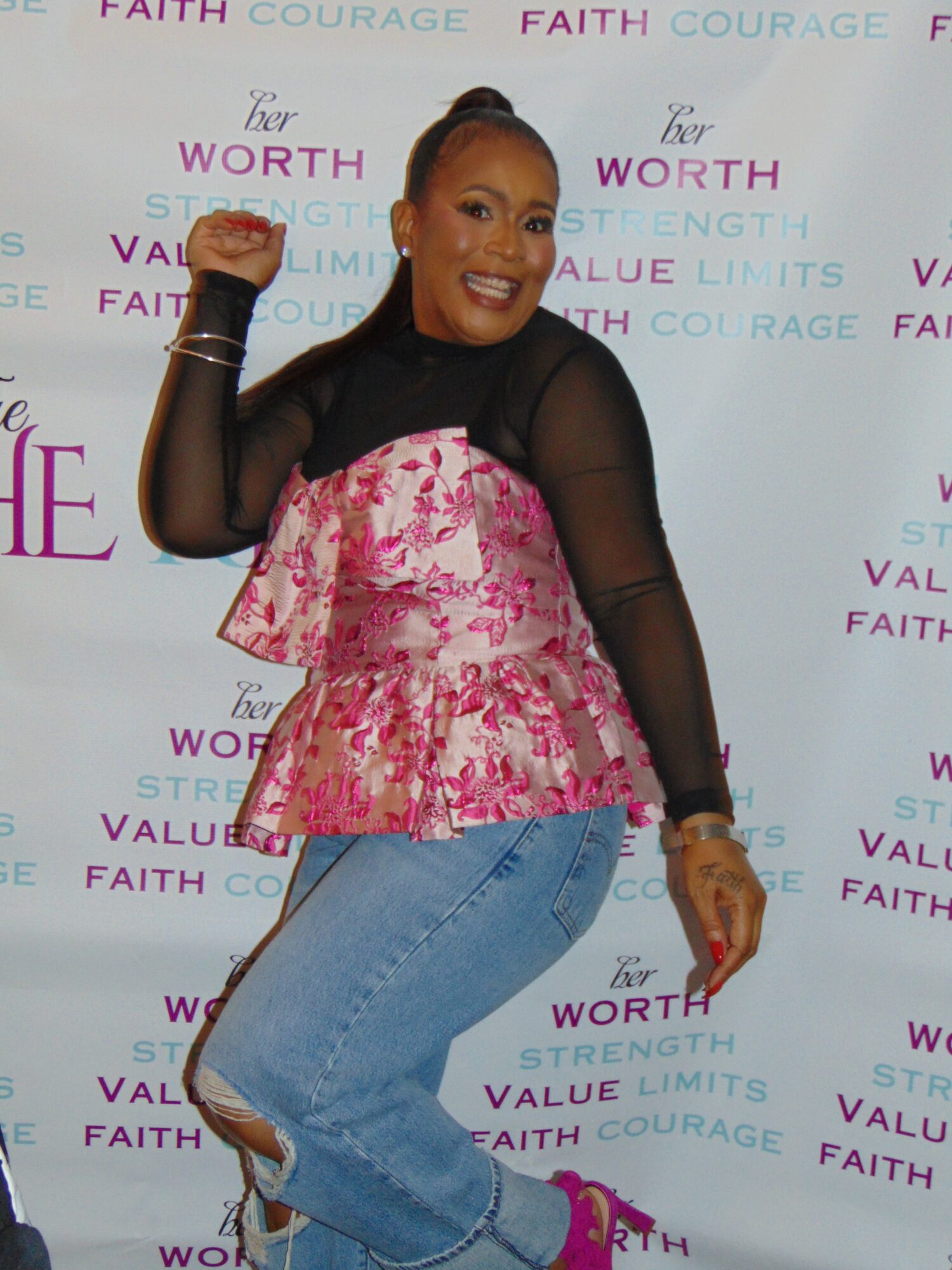 Smiling woman in black and pink floral top, blue jeans, and pink shoes, posing with one arm raised, against a patterned background.