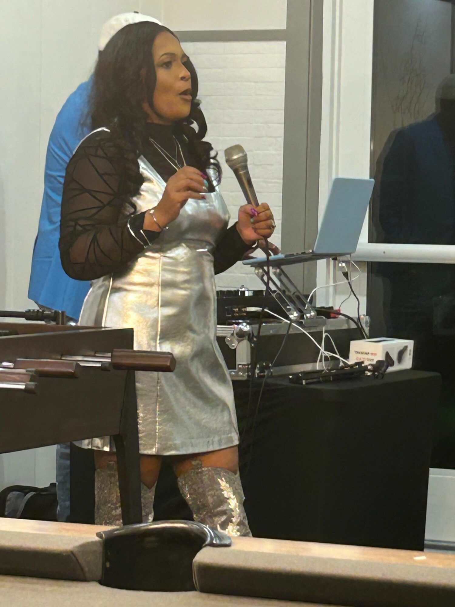 Woman standing indoors, holding a microphone, wearing a shiny silver dress and black jacket, with a window and equipment behind her.