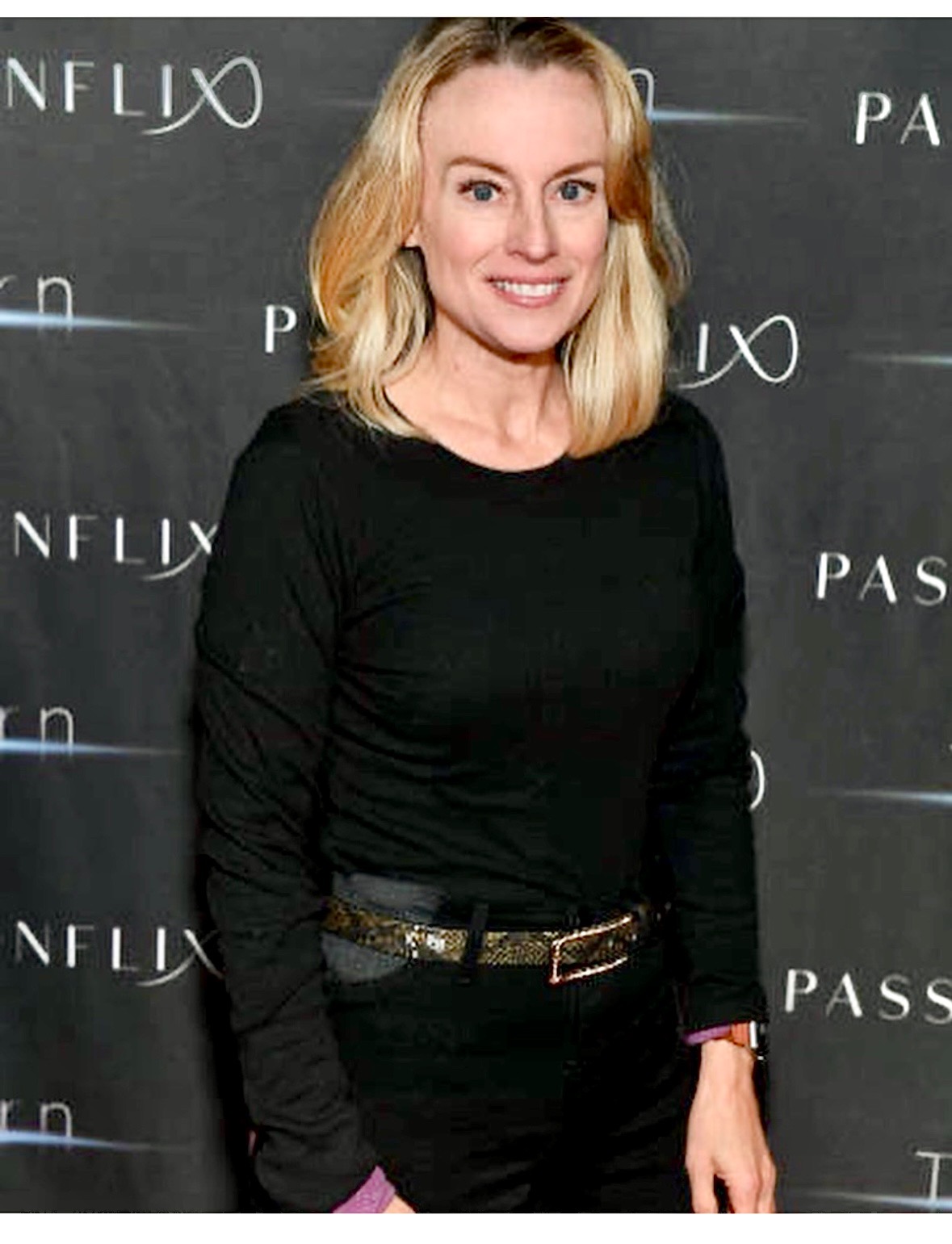 Woman with blonde hair wearing a black long-sleeve top and black pants, standing in front of a backdrop with Netflix and Pass logos.