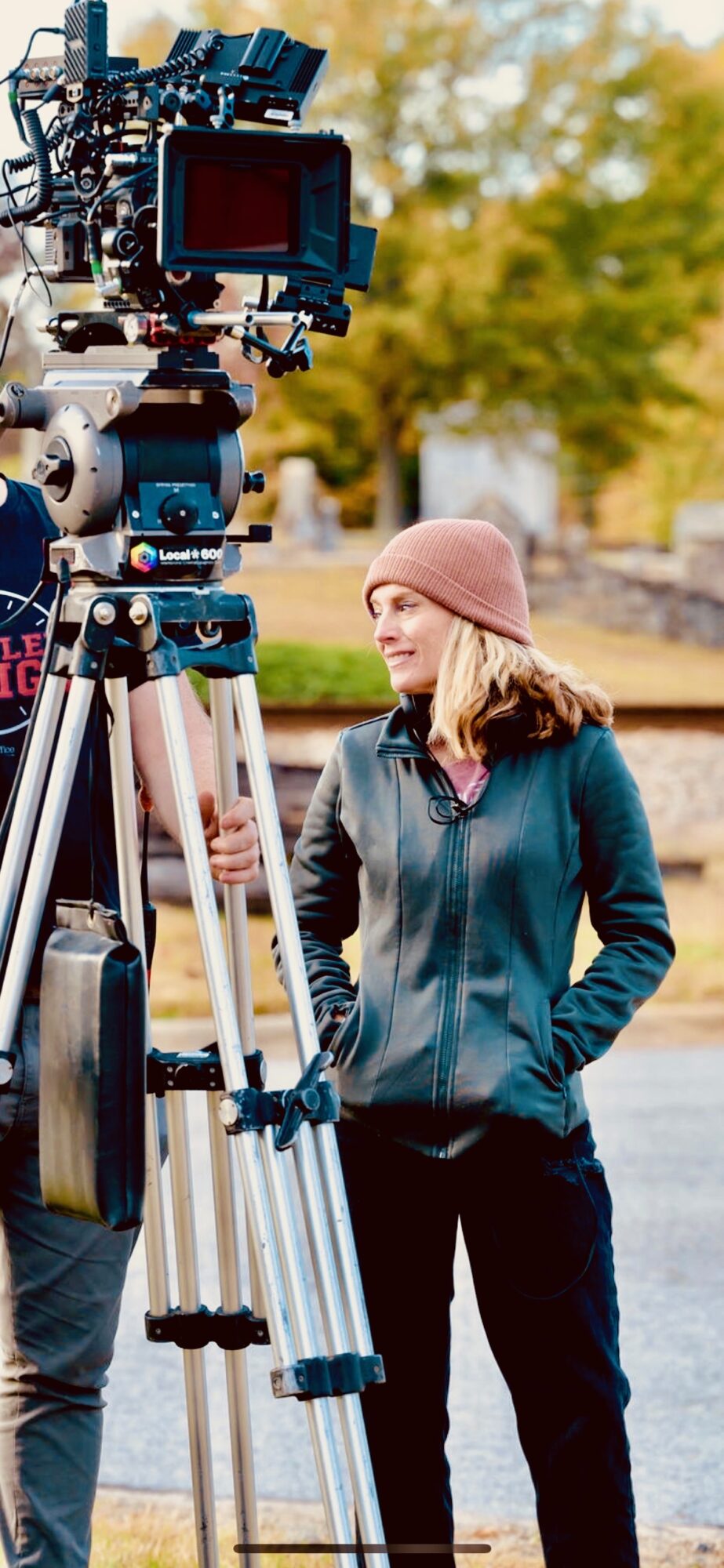 Woman in pink hat and blue jacket standing outdoors near water, being filmed with professional camera equipment.