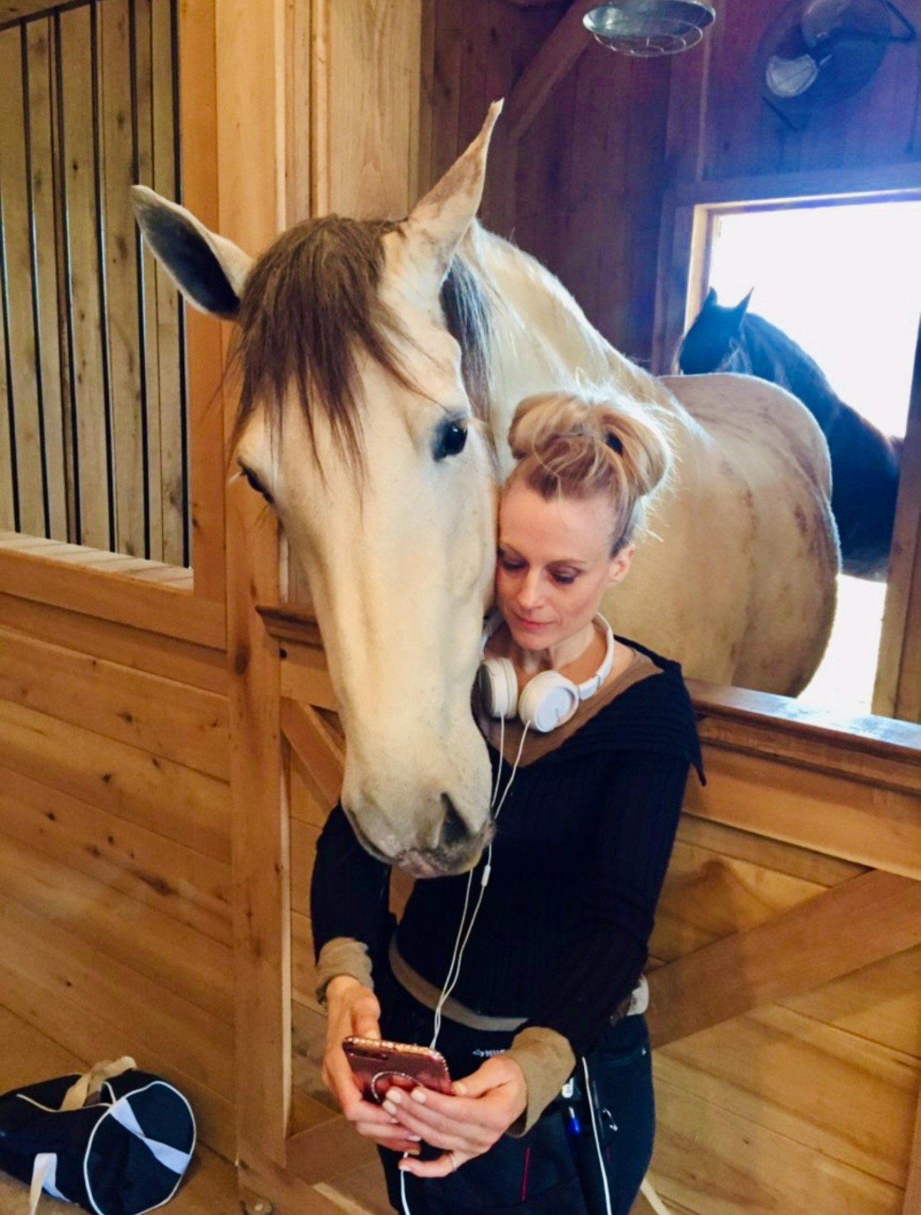 Woman with headphones holding a phone, leaning against a horse's head inside a wooden stable, with another horse visible outside.