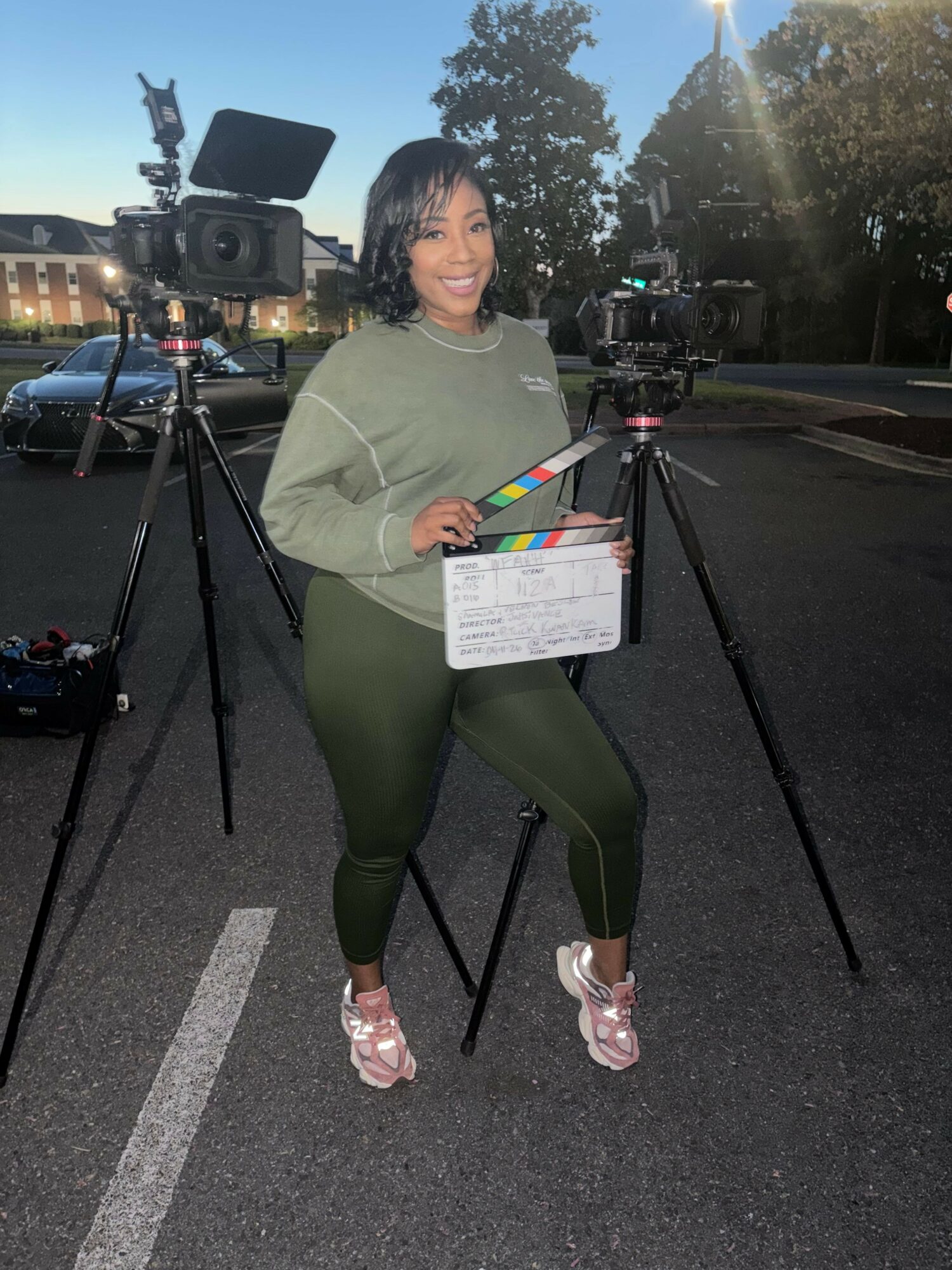 Woman holding a clapperboard standing on a street with camera equipment, trees, and buildings in the background.