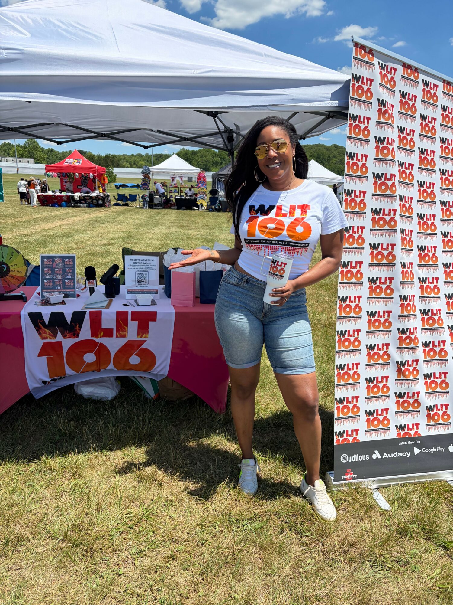 Woman standing outdoors next to a table with promotional signs, wearing a white T-shirt and shorts, smiling.