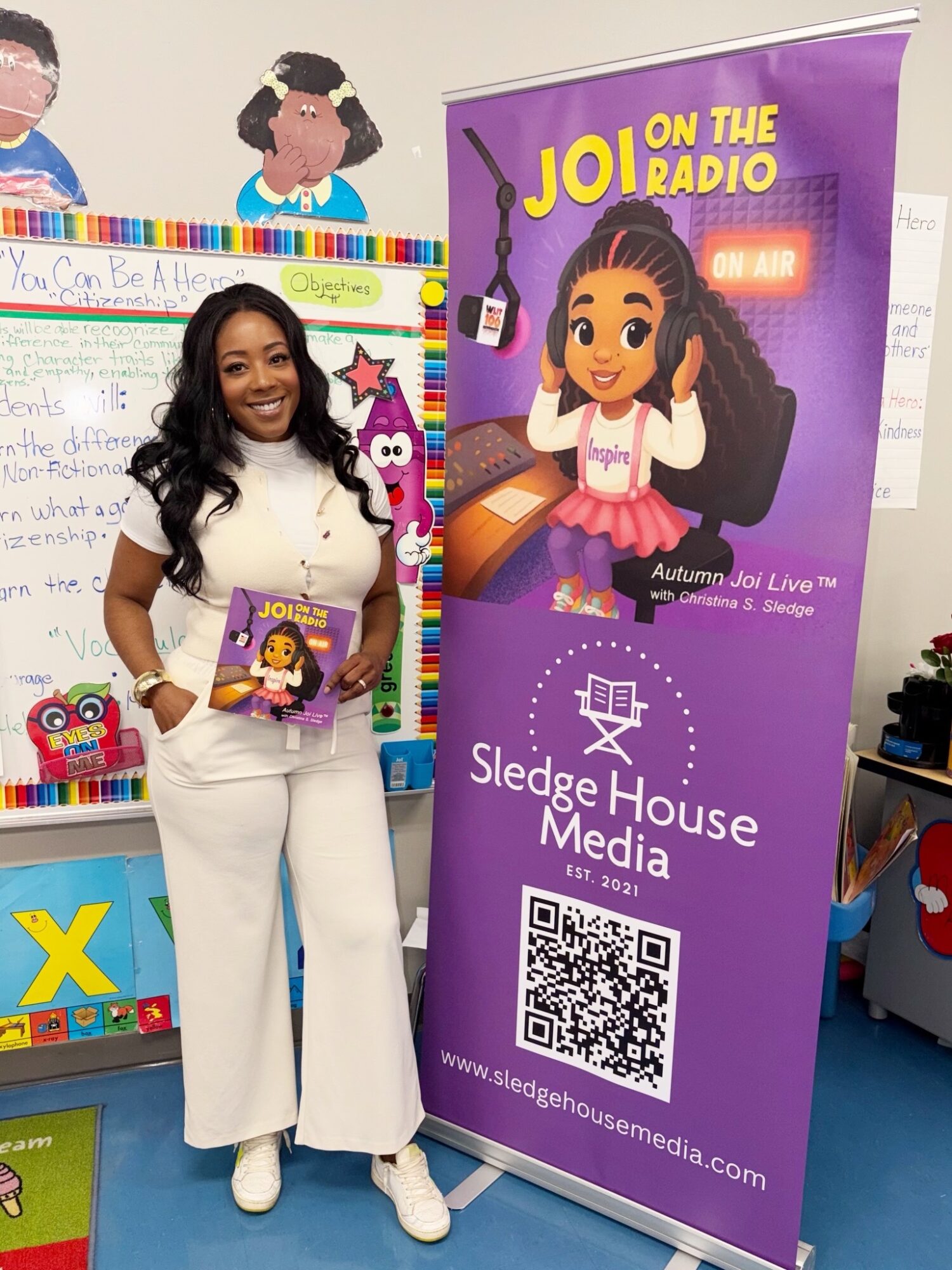 Woman standing indoors holding a book, smiling, next to a purple Sledge House Media banner with cartoon girl and QR code.