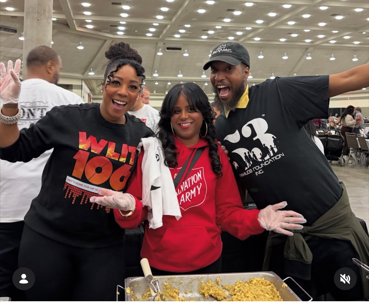 Three smiling people standing together indoors, with food in front of them, arms around each other, celebrating.