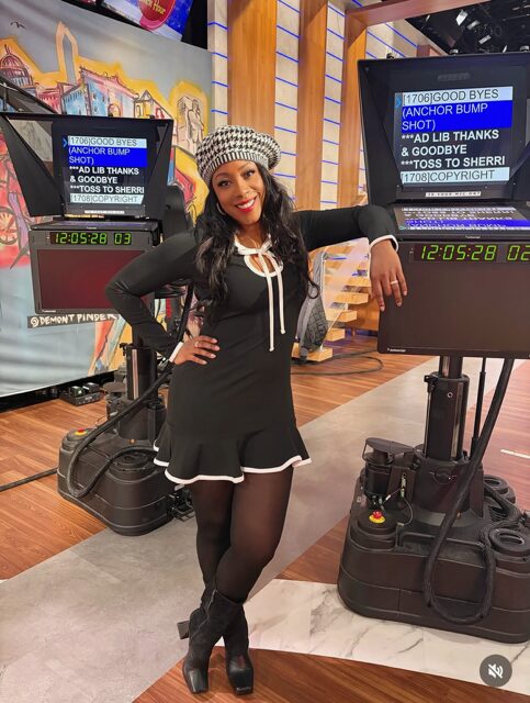 Woman in black outfit and checkered hat smiling, standing in a TV studio with cameras and screens around her.
