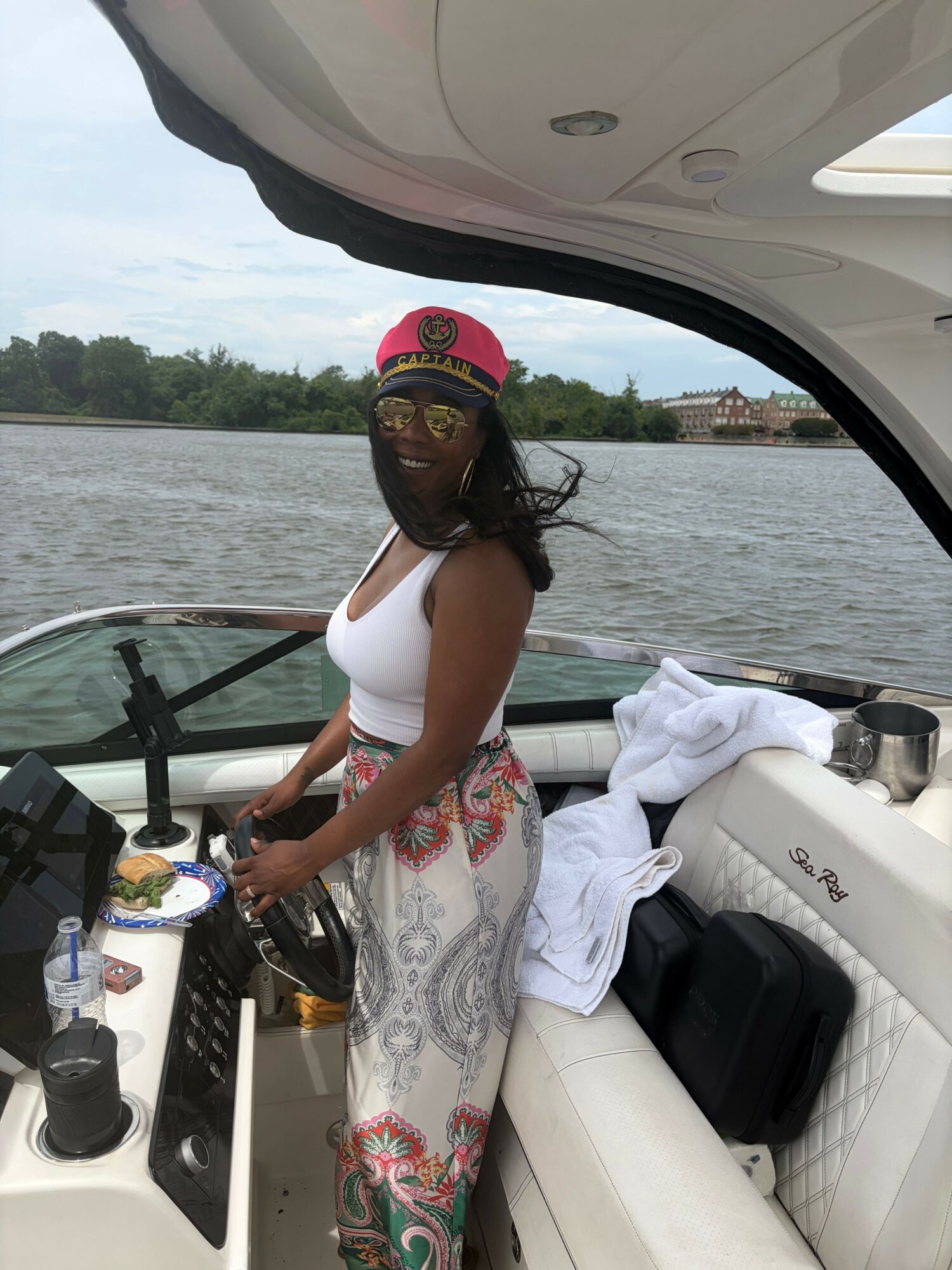 Woman standing on boat, holding a drink, with water and trees in background.