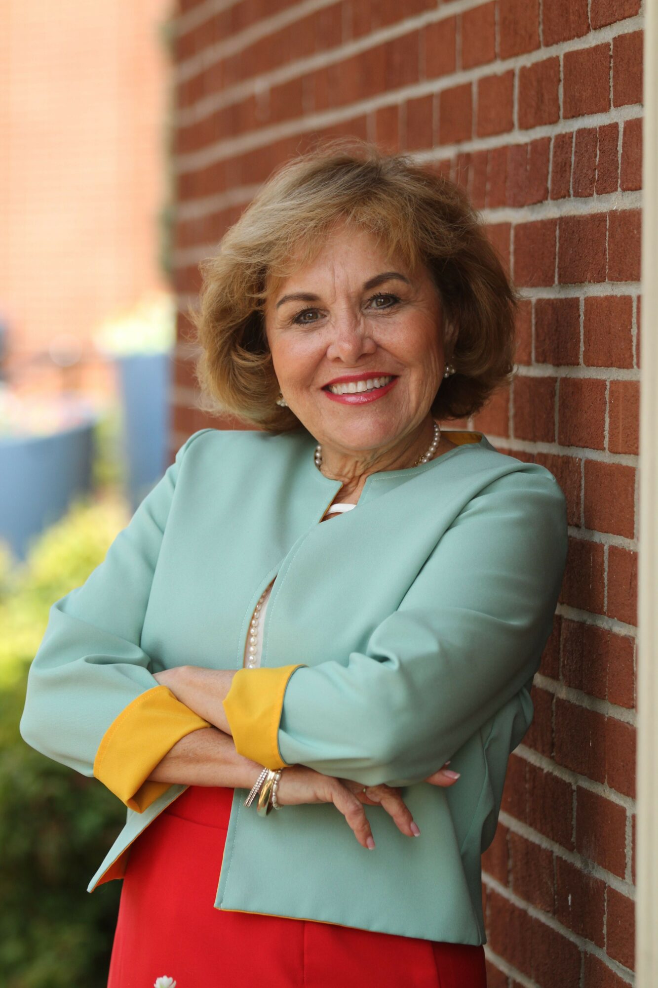 Woman with blonde hair smiling, wearing a light blue blazer with yellow cuffs, standing outdoors against a brick wall.