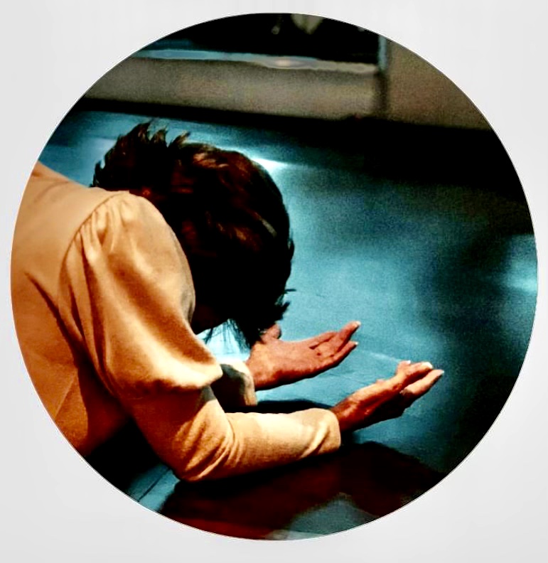 Person with dark hair leaning over a surface, hands extended downward, in a circular frame.