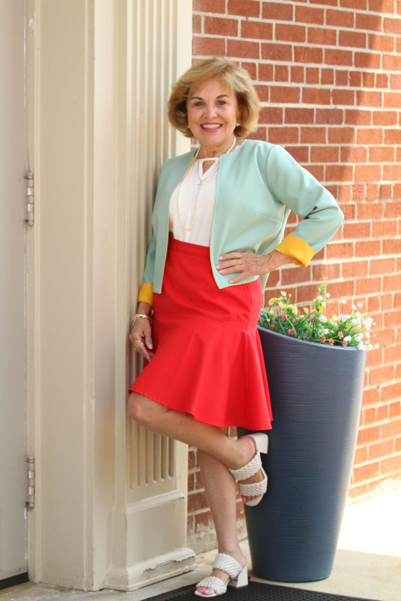 Woman standing outside near a brick wall, wearing a red skirt, light blue jacket, and white blouse, smiling.
