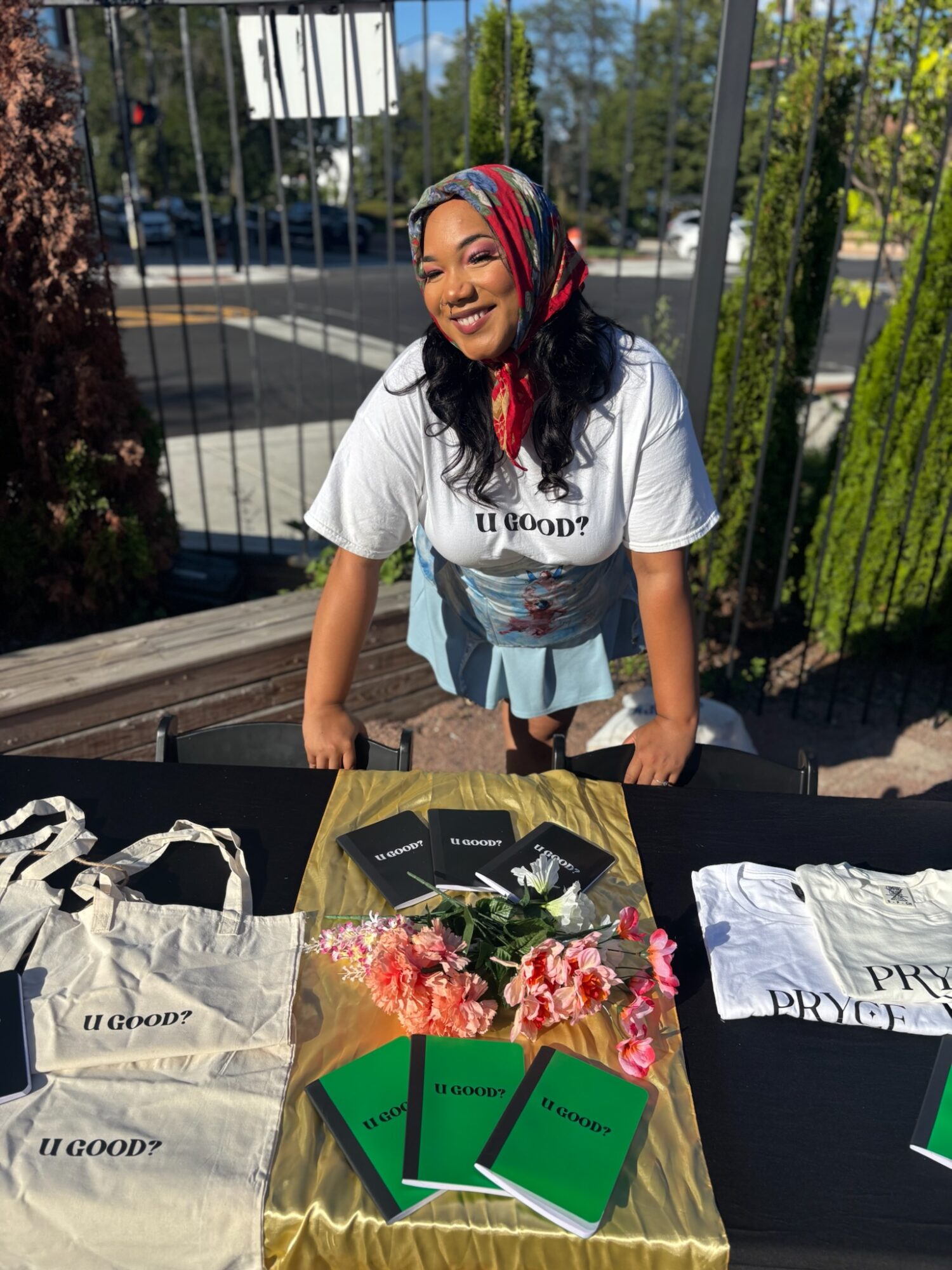 Young woman with a colorful headscarf smiling, standing behind a table with books, flowers, and bags outdoors.