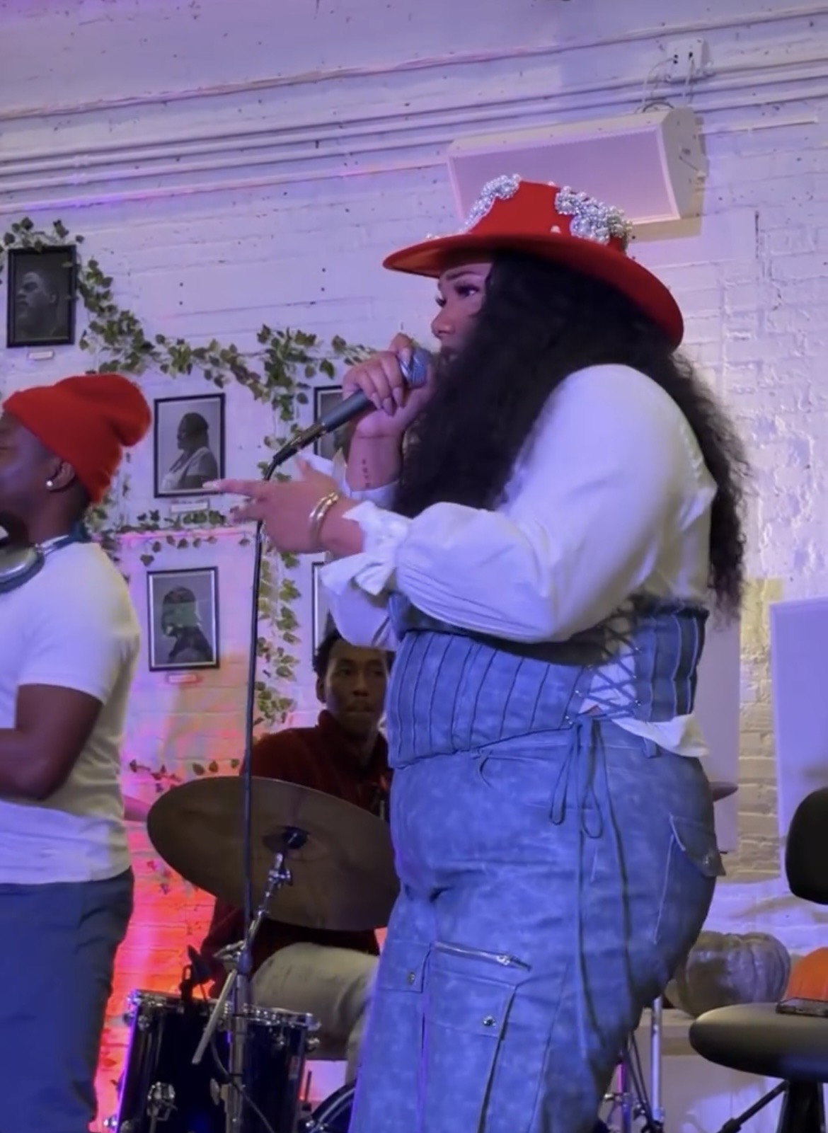 A woman wearing a red hat with decorations, white blouse, and blue overalls singing into a microphone at an indoor event.