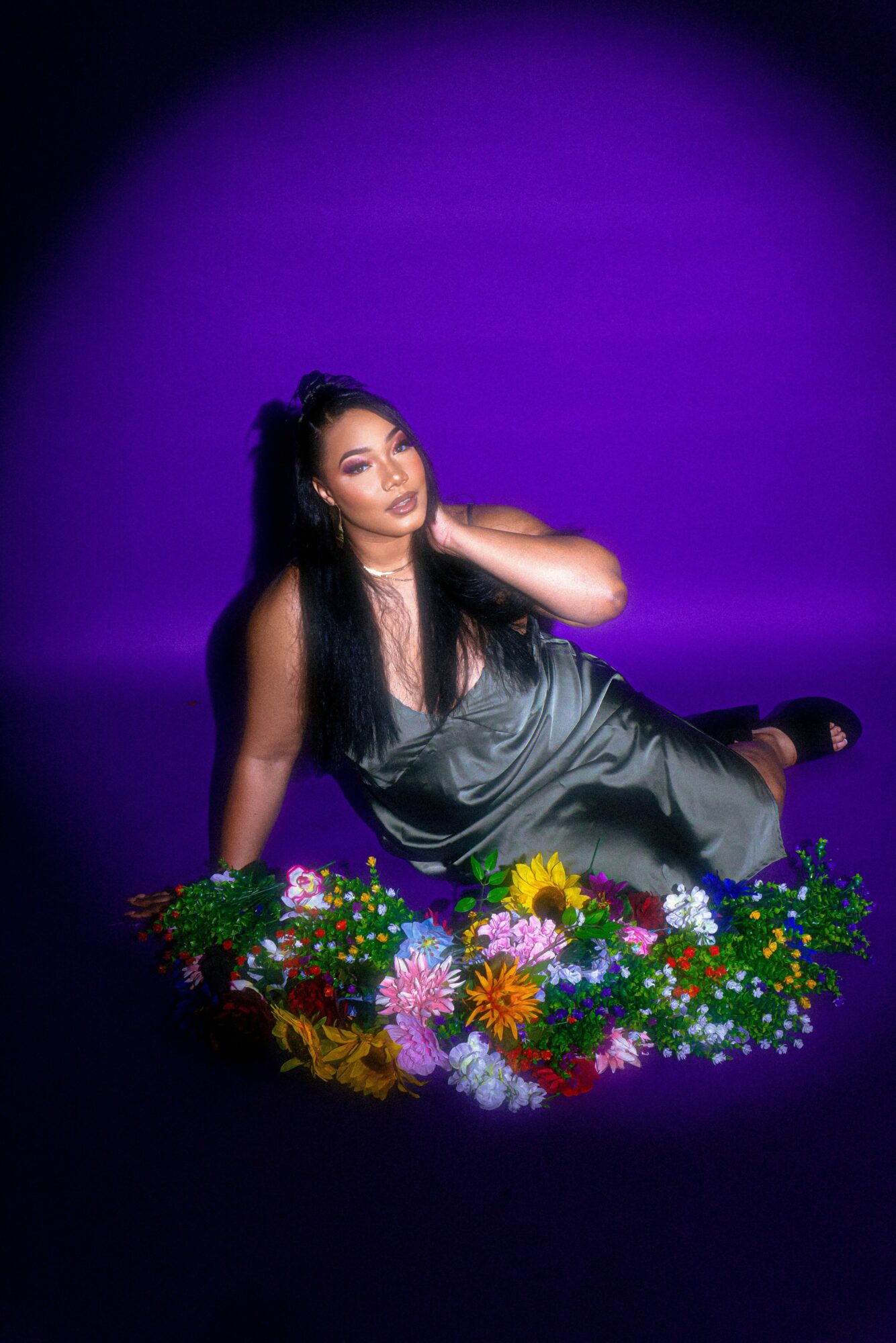 Woman lying on the floor surrounded by colorful flowers, with a purple background, wearing a shiny dress, touching her hair.
