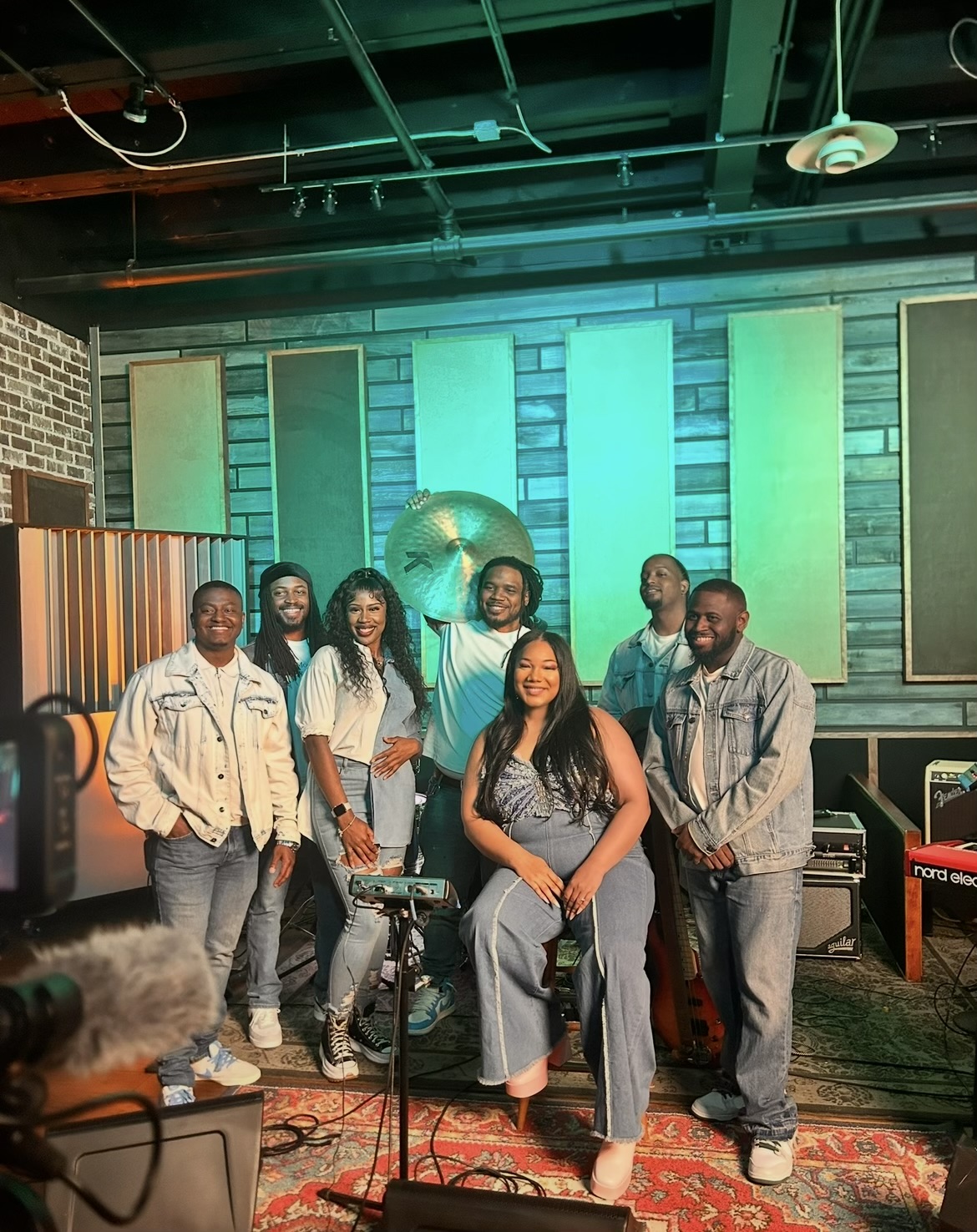Group of seven people posing in a music studio with colorful acoustic panels and musical equipment.