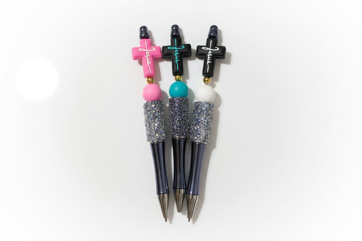 Three pens with cross-shaped toppers in pink, teal, and black, each with glittery silver grips and black bodies.