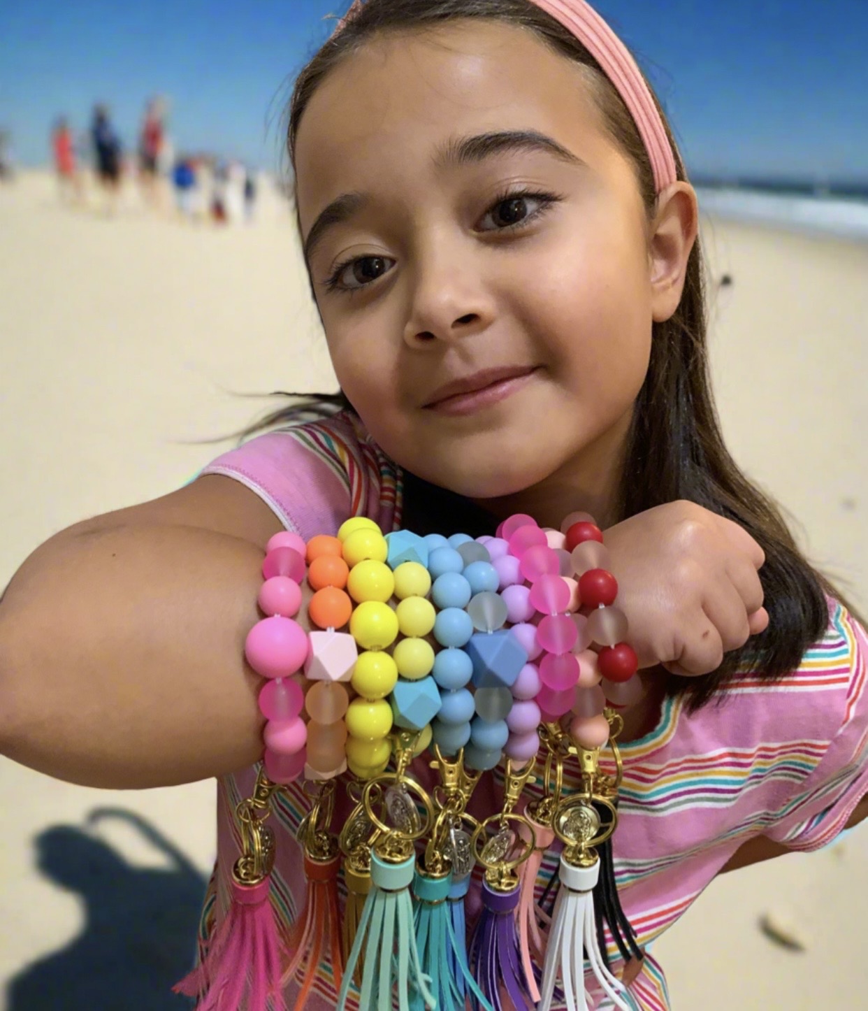 Young girl with colorful beaded bracelets on her arm at the beach, smiling, with sand and people in the background.