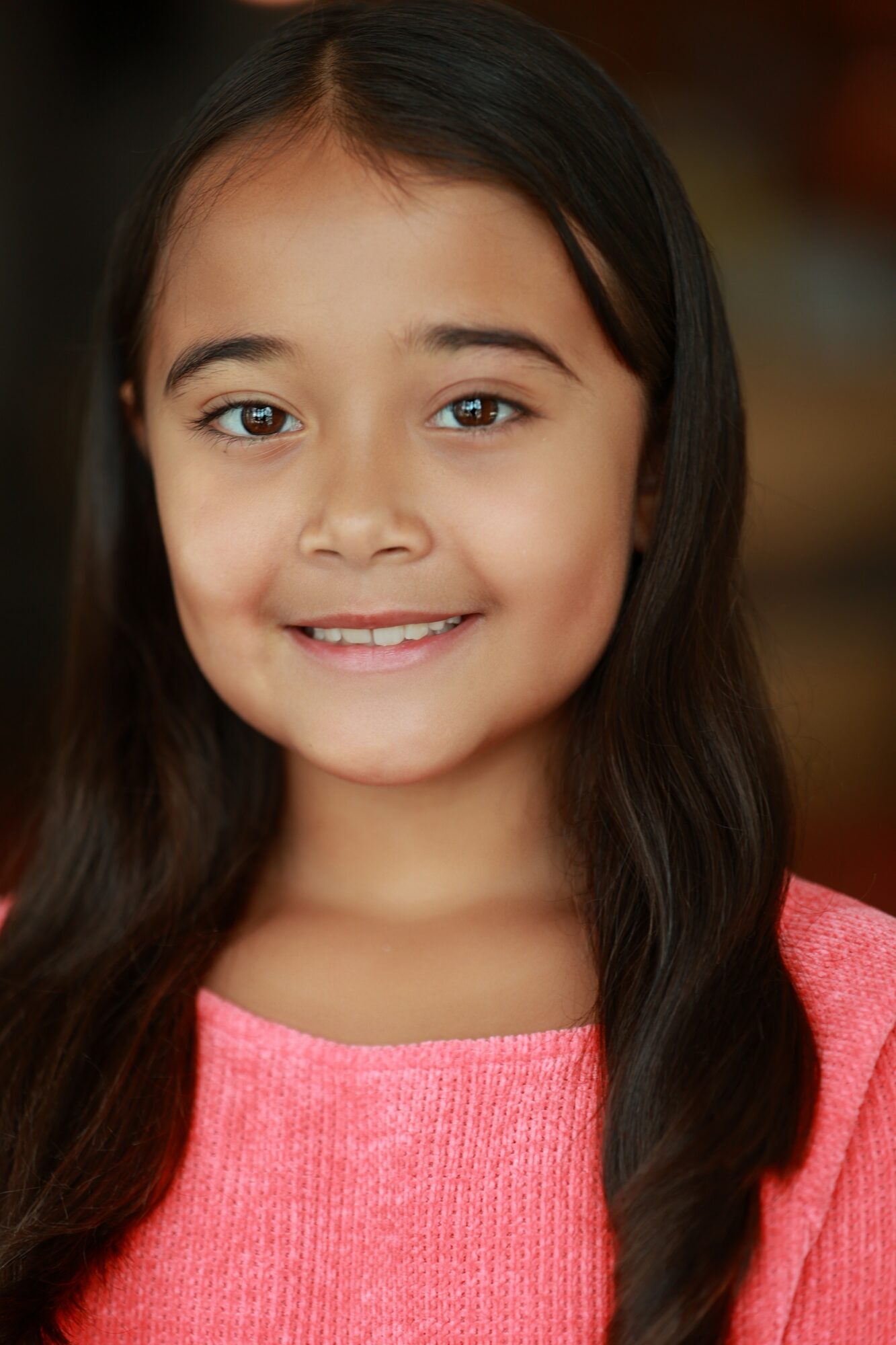 Young girl with long dark hair, smiling, wearing a pink top, facing forward.