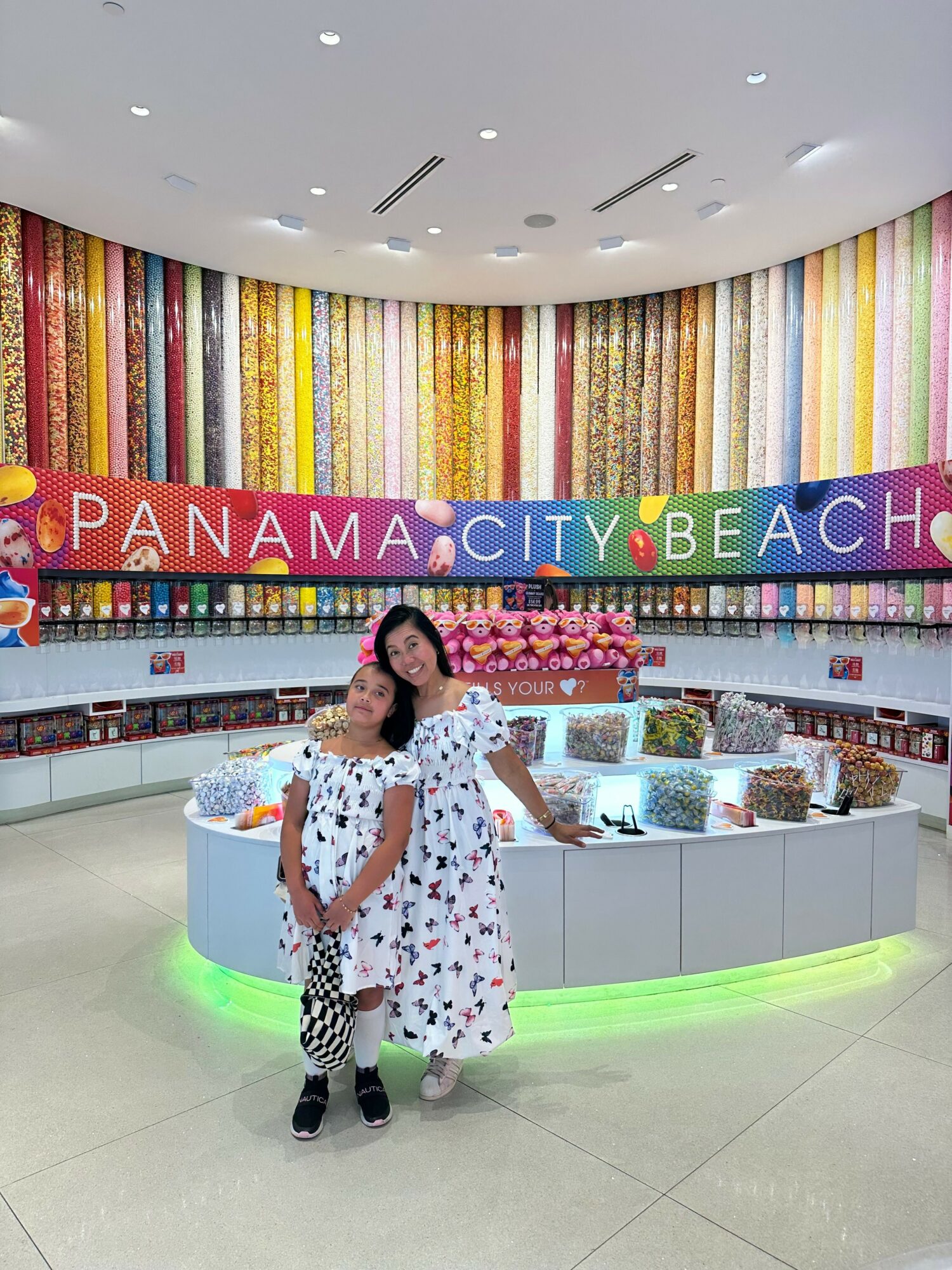 Woman in a white dress with black patterns stands near a display table with colorful items, in a brightly lit indoor space.