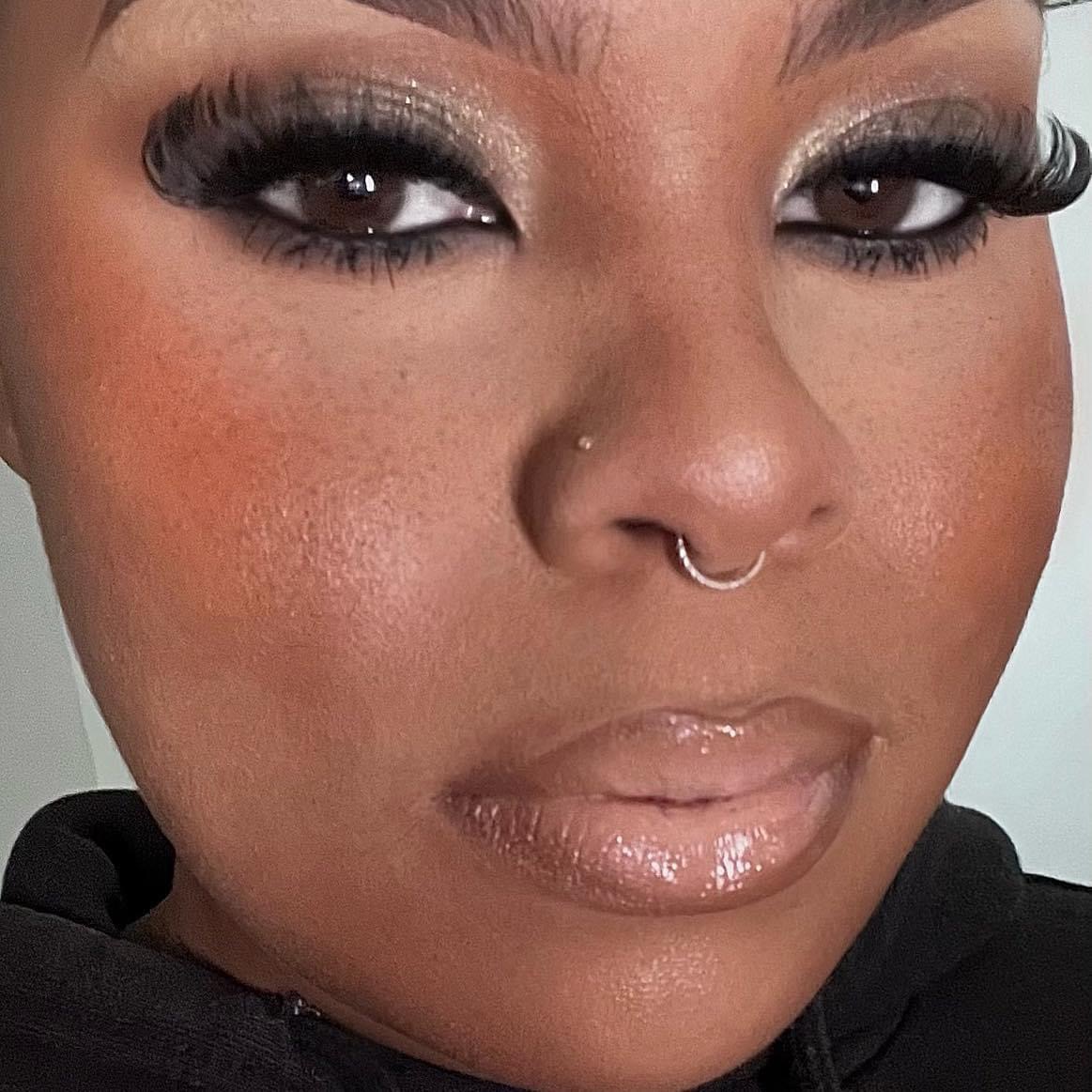 Close-up of a person's face with makeup, nose piercing, and glossy lips, looking directly at the camera.