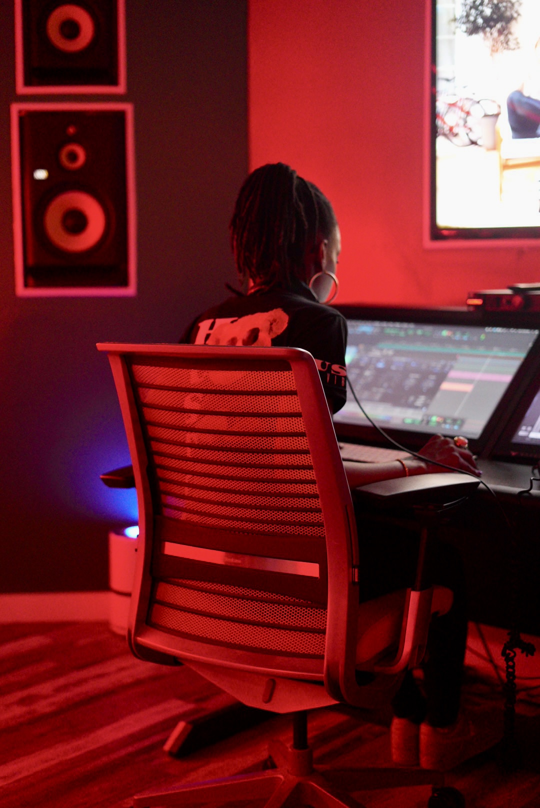 Person with dreadlocks working at a computer in a dimly lit room with red lighting, speakers on the wall, and a window showing outside.