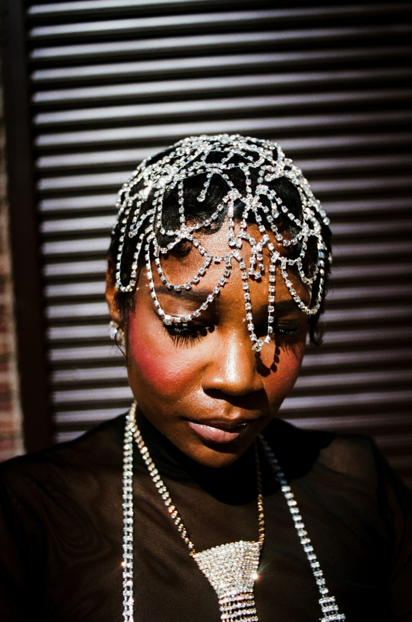 Woman with intricate pearl headpiece and necklace, closed eyes, dark background with horizontal lines.