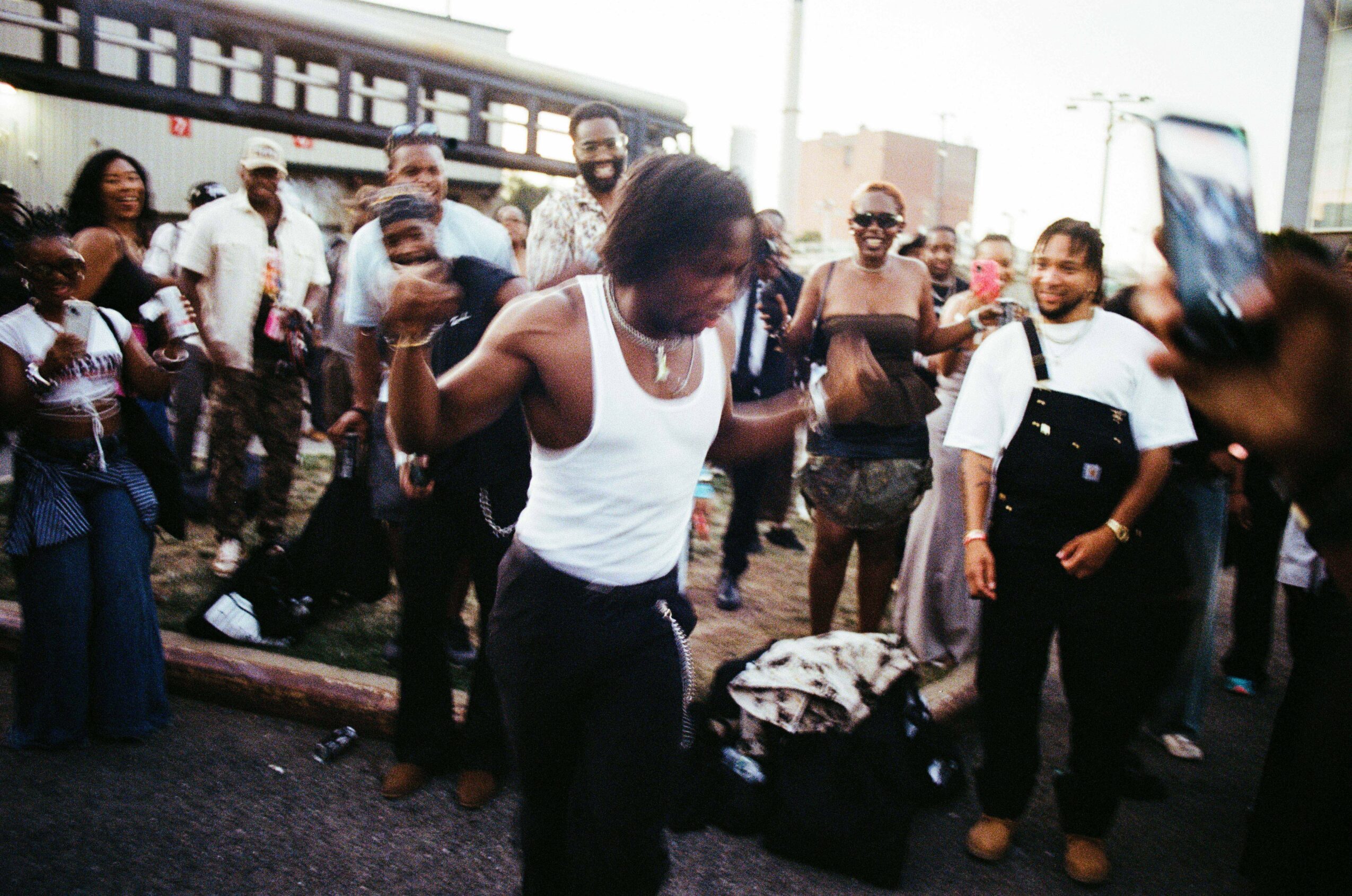 Group of people gathered outdoors, some dancing and taking photos, with a woman in white tank top in center.