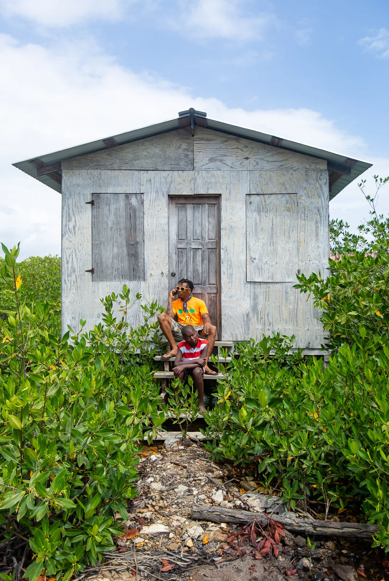 Child sitting on a bench in front of a weathered wooden house surrounded by green plants, under a partly cloudy sky.