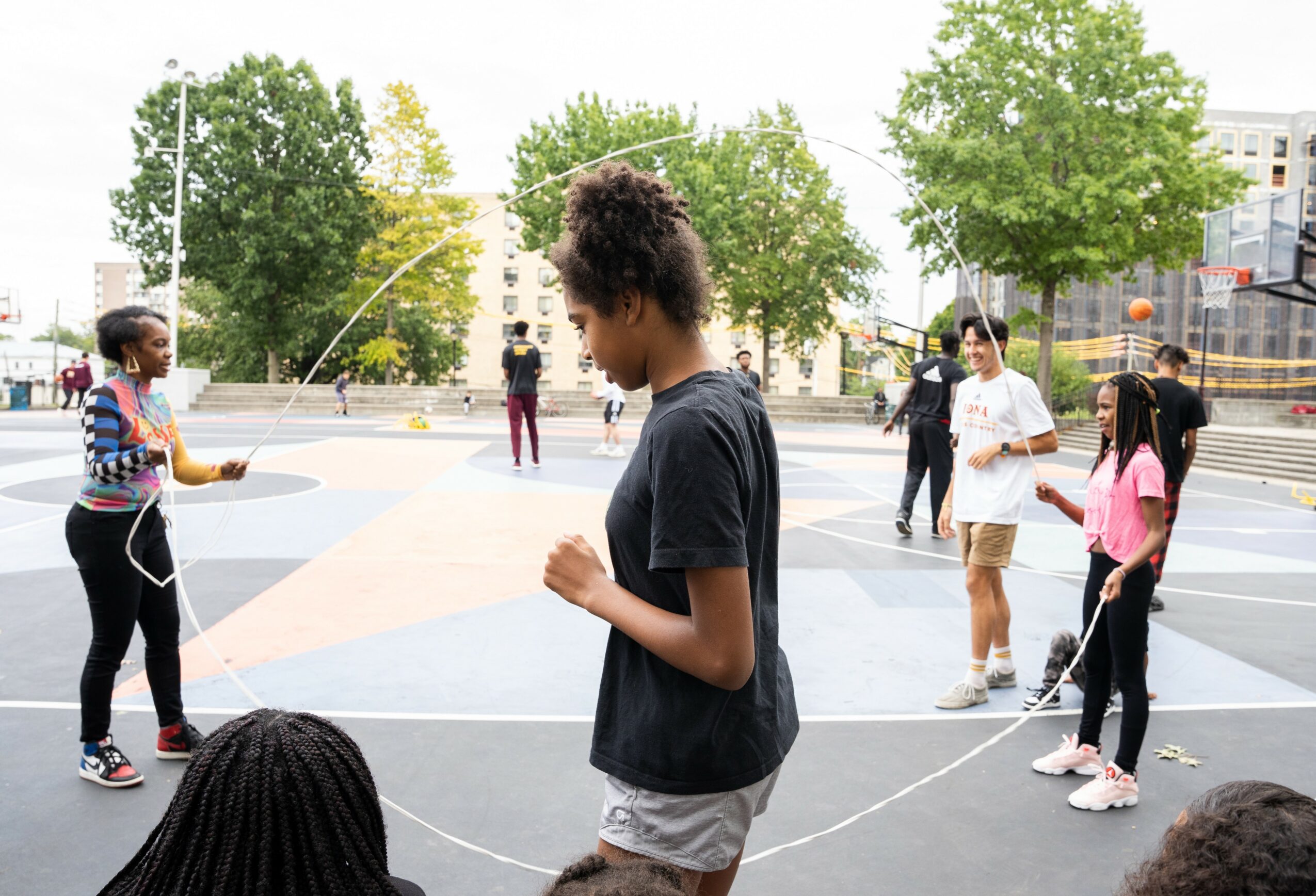 Group of children playing with jump ropes outdoors in a park with trees and buildings in the background.