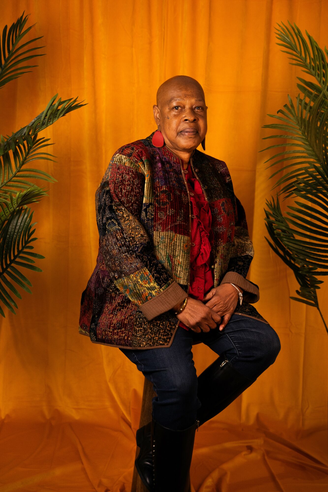 Person seated in front of a yellow curtain, wearing a patterned jacket and red shirt, with green plants on sides.
